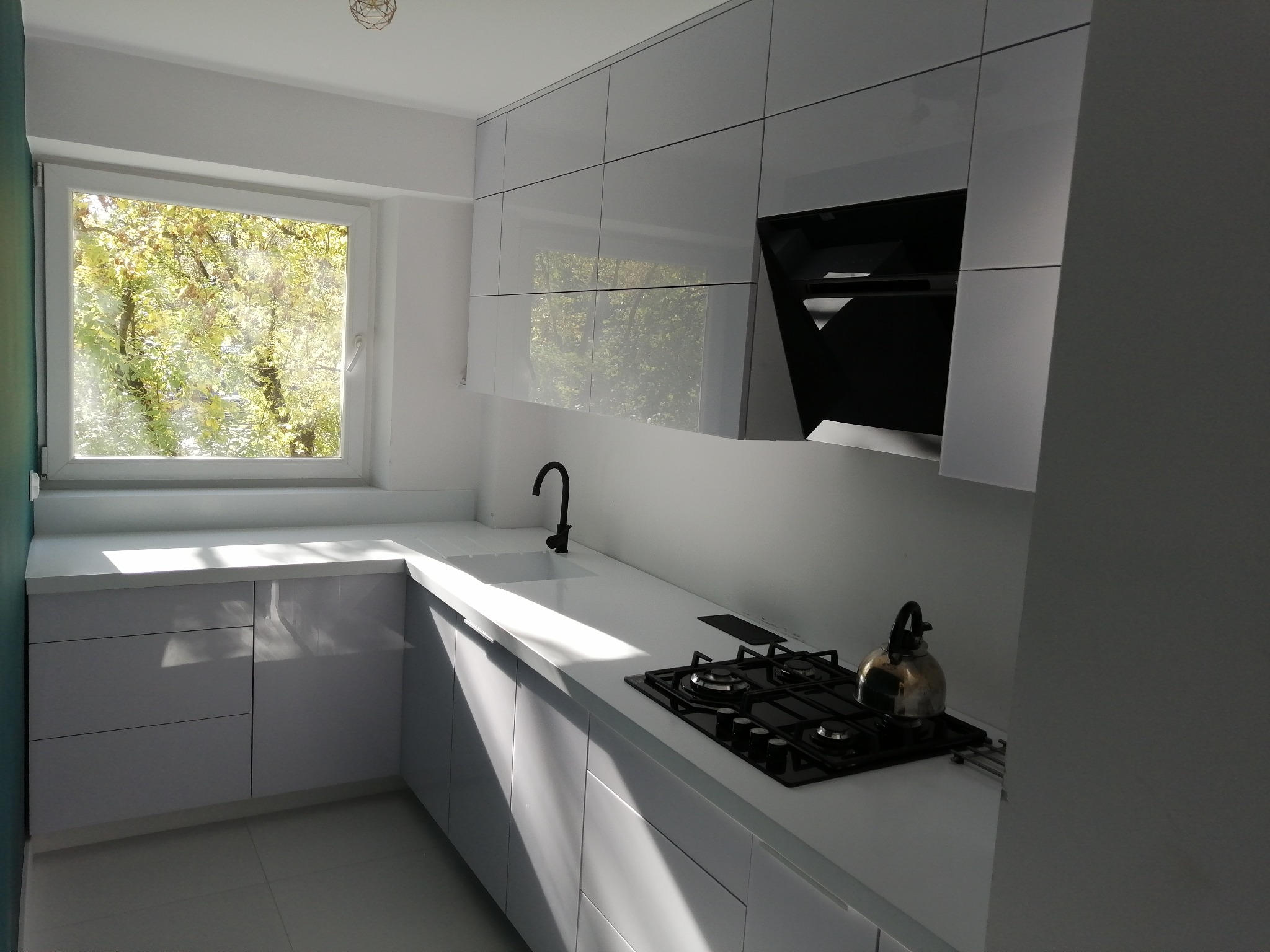 Nowoczesna kuchnia z białymi, lakierowanymi frontami, czarną armaturą i płytą gazową, oświetlona naturalnym światłem z okna z widokiem na drzewa.