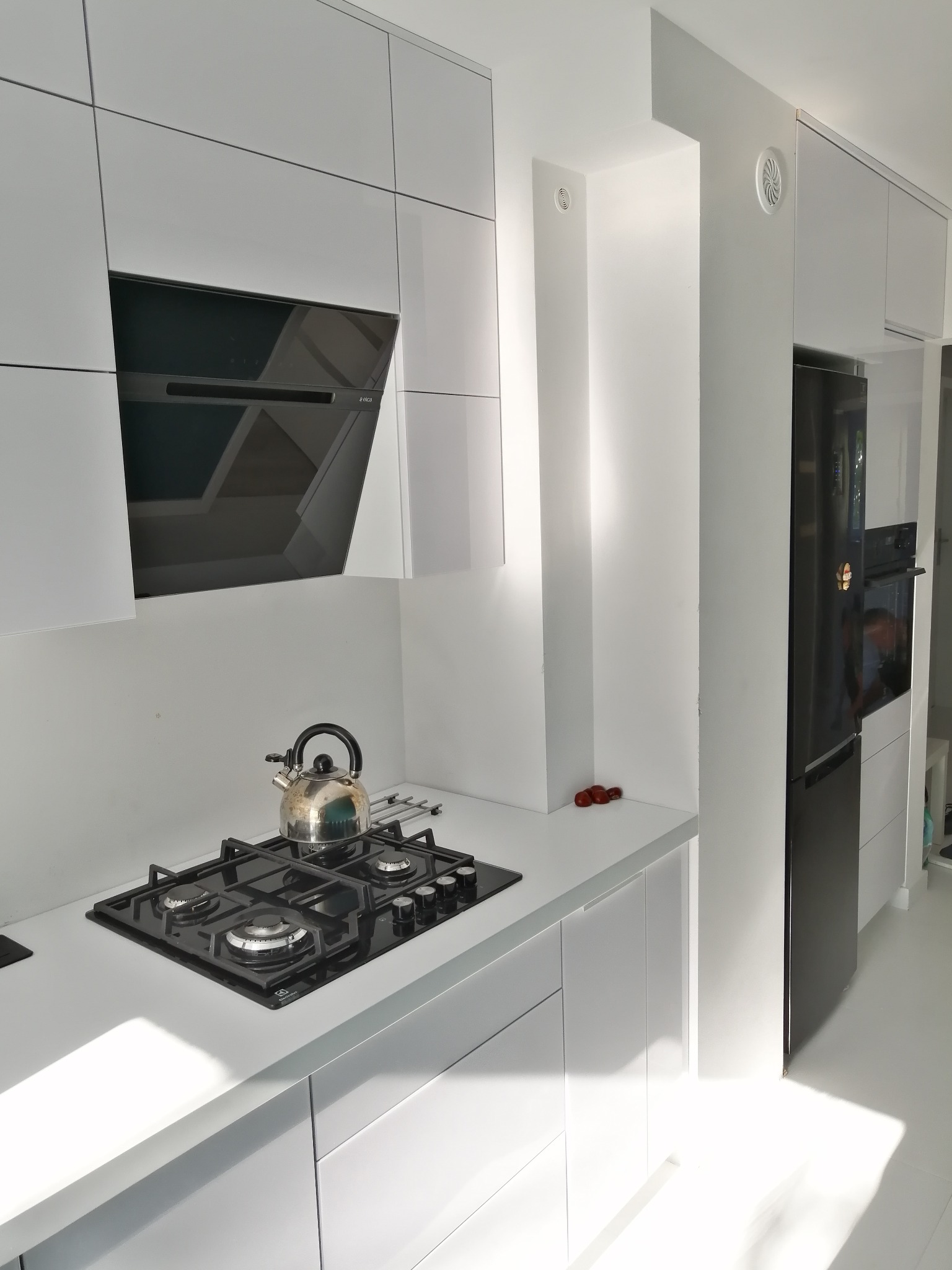Nowoczesna, minimalistyczna kuchnia z białymi, lakierowanymi frontami, czarną płytą gazową z czajnikiem i czarnym okapem nad nią, oraz czarną lodówką w zabudowie.