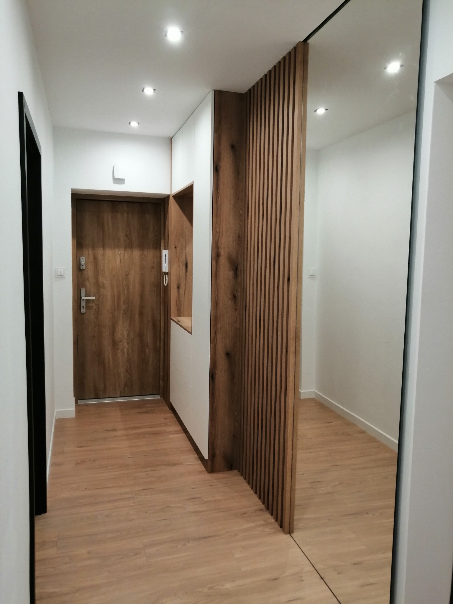 Nowoczesny przedpokój z drewnianą zabudową, białą szafą z otwartą półką, lustrem na ścianie i drewnianą podłogą.