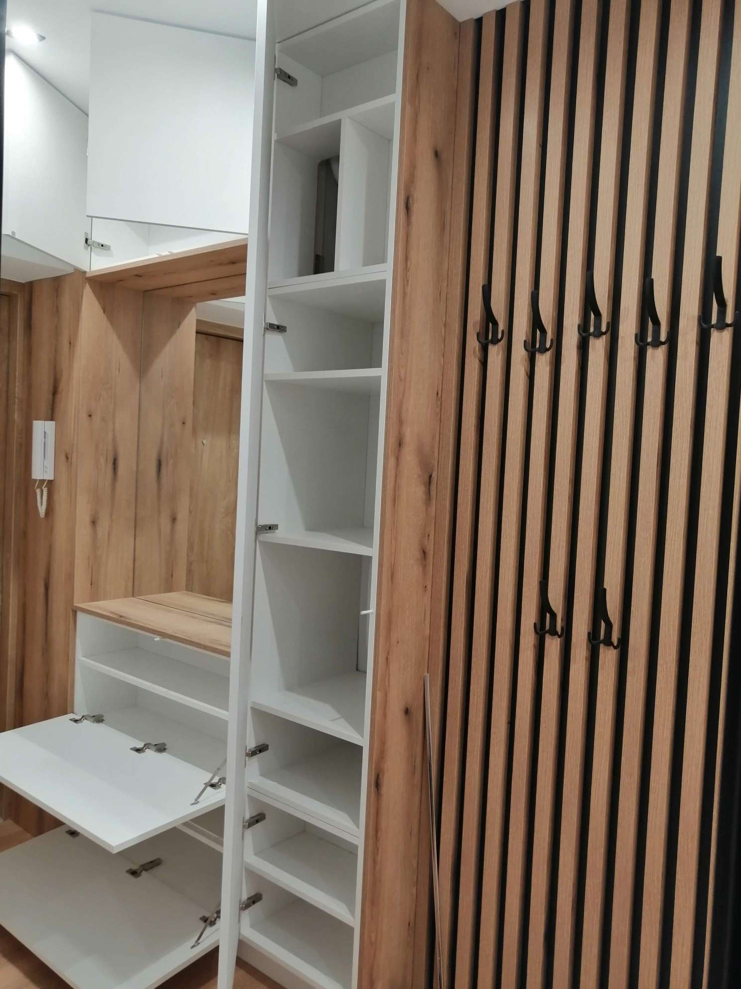 Wnętrze przedpokoju z białą szafą z otwartymi półkami i drewnianymi elementami, w tle ściana z drewnianych listew z czarnymi haczykami.