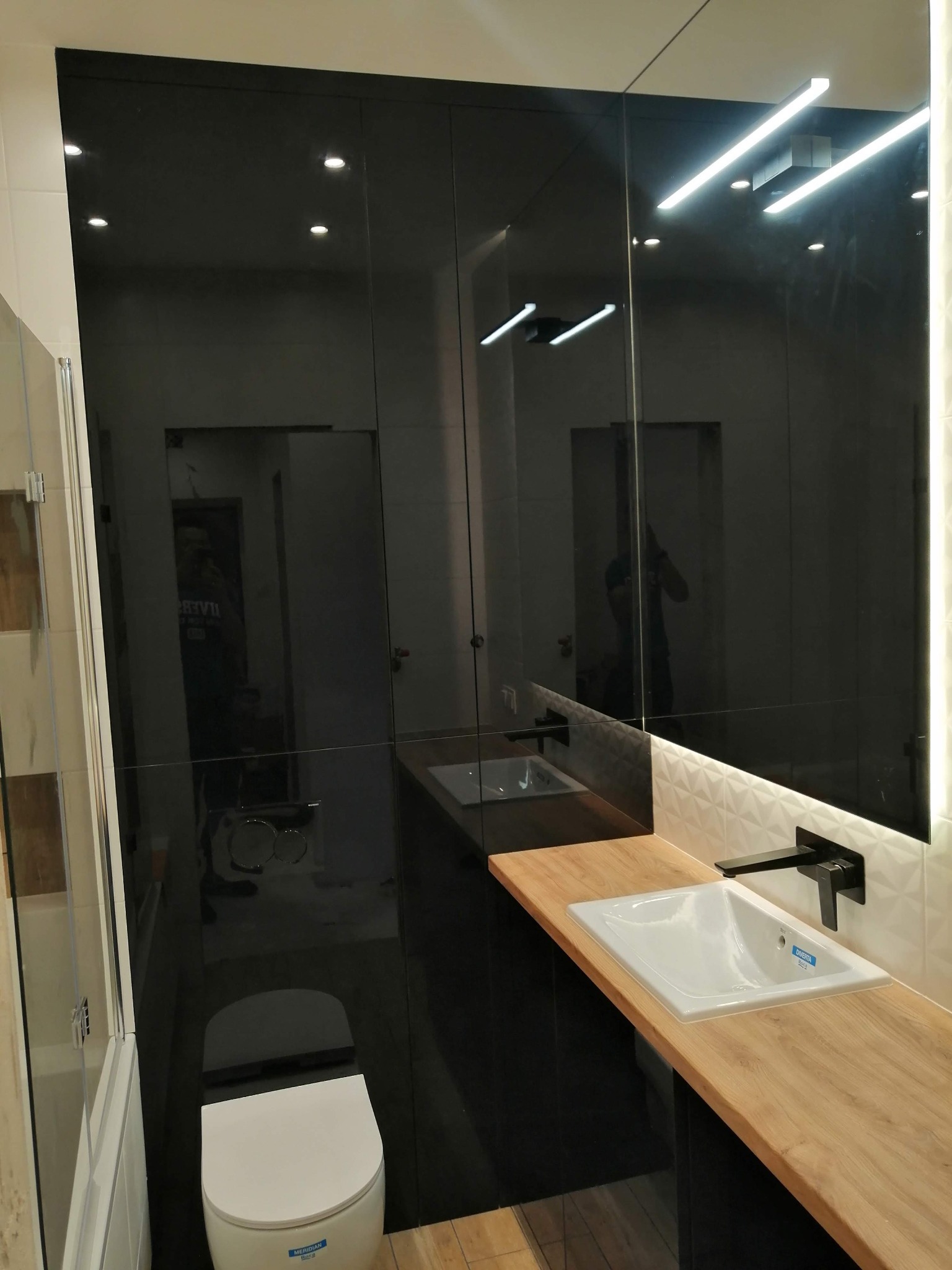 Nowoczesna łazienka z czarnymi, lakierowanymi szafkami, drewnianym blatem pod umywalką i białym sedesem. Oświetlenie LED podkreśla geometryczne wzory na ścianie.