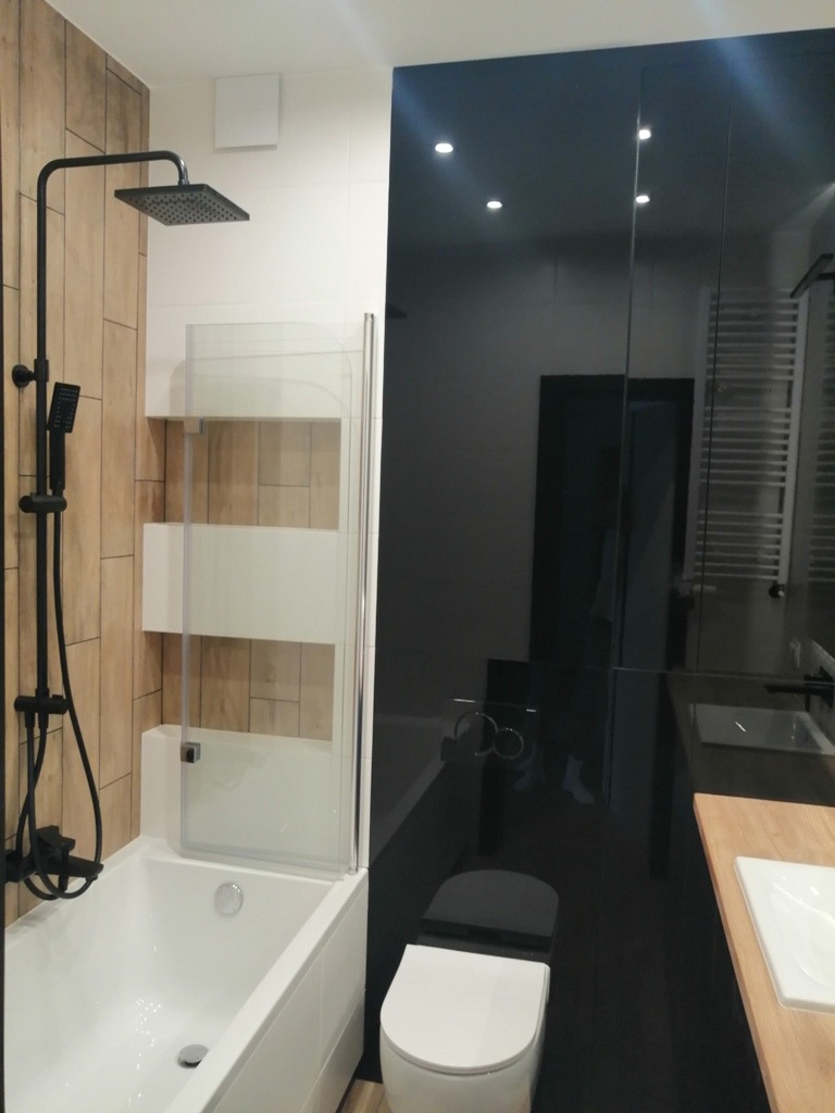 Nowoczesna łazienka z wanną, czarną armaturą i błyszczącymi czarnymi panelami ściennymi, uzupełniona drewnianymi akcentami.