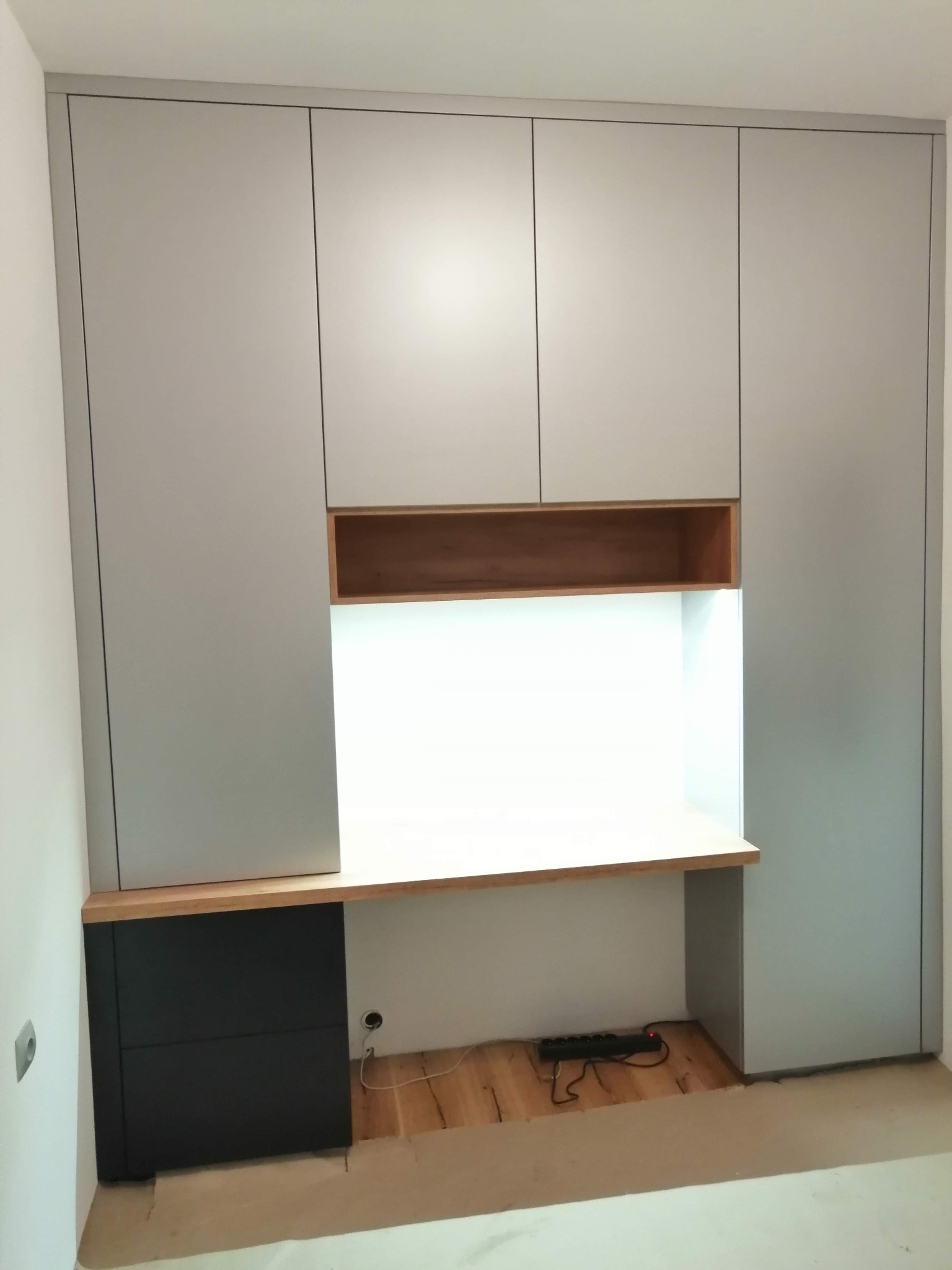 Szara zabudowa meblowa z biurkiem i szafkami, drewniane elementy wykończeniowe, widoczny przedłużacz na podłodze w trakcie montażu.
