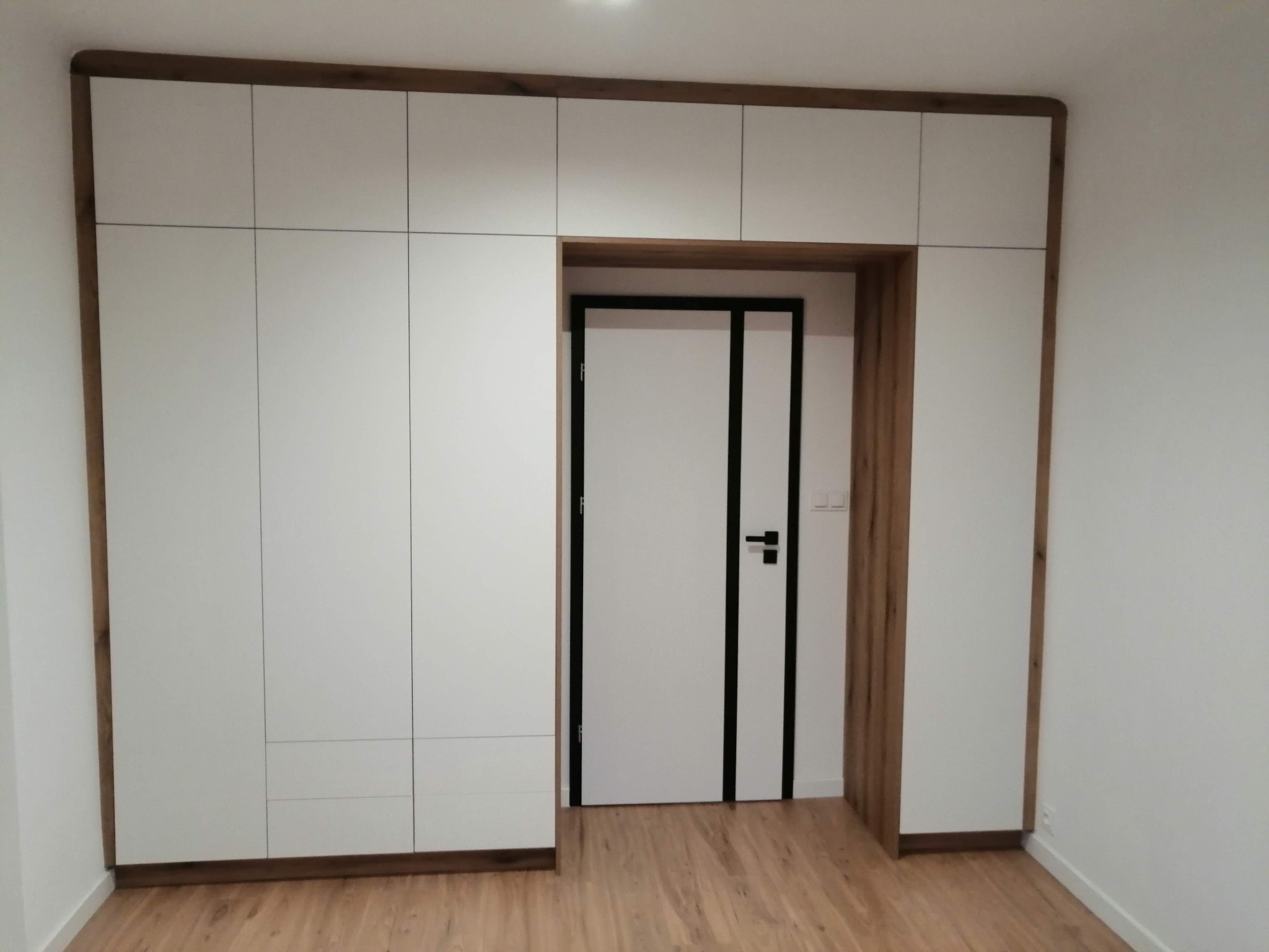 Biała szafa wnękowa z drewnianą ramą okalającą również drzwi wejściowe, tworząc spójny wizualnie układ zabudowy.