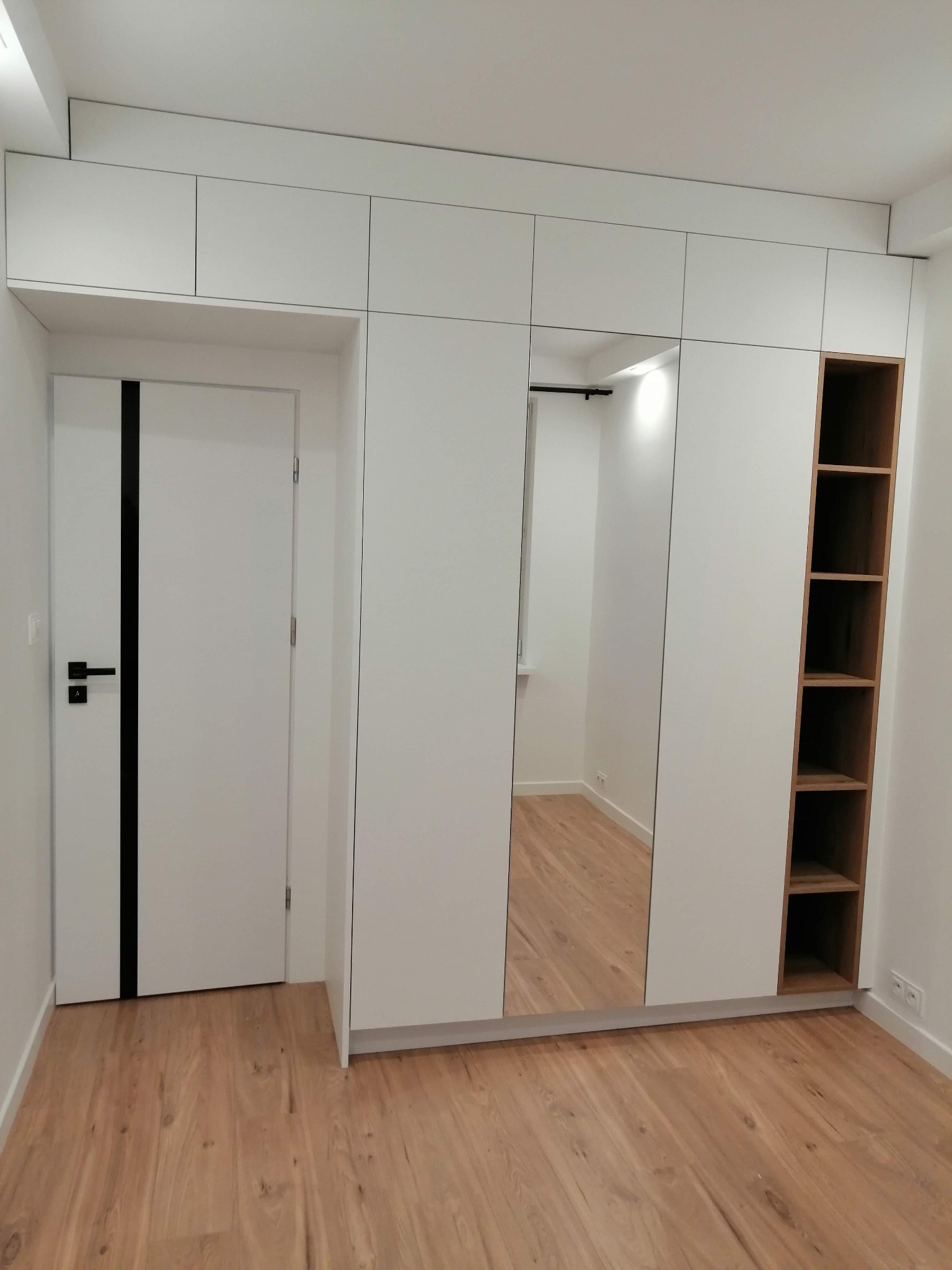 Biała szafa wnękowa z lustrem i otwartymi półkami z jasnego drewna, dopasowana do białych drzwi z czarnym akcentem w nowoczesnym wnętrzu z drewnianą podłogą.