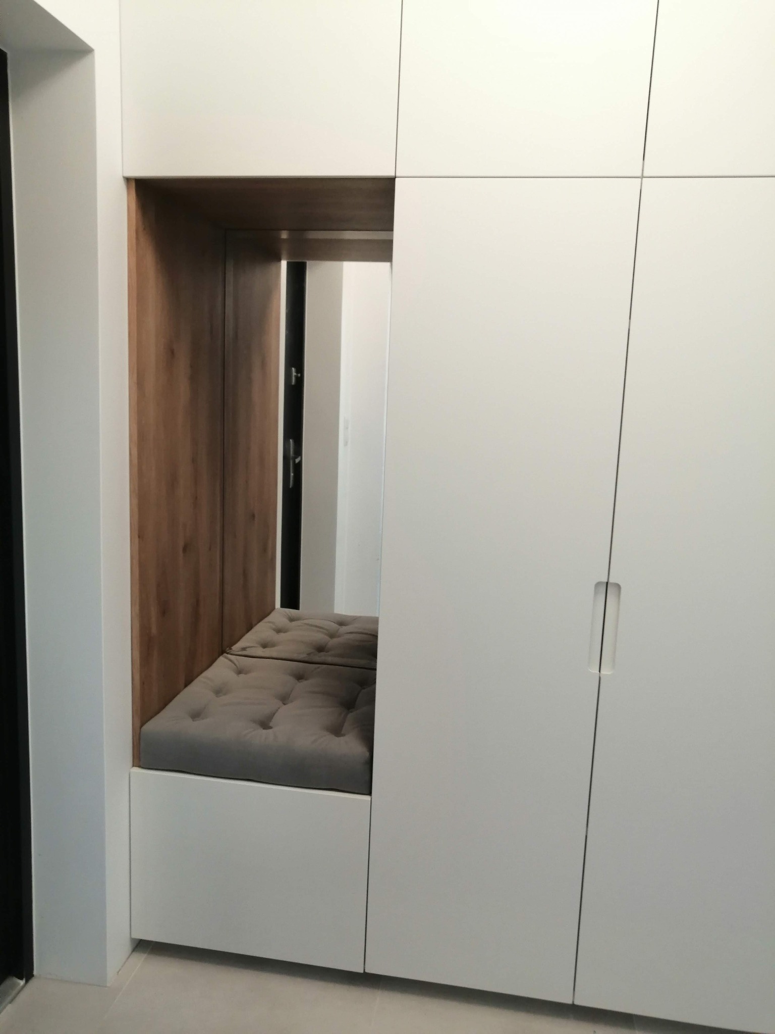 Biała szafa wnękowa z siedziskiem z szarej tapicerki z guzikami, drewniane wykończenie wnęki, minimalistyczny design.