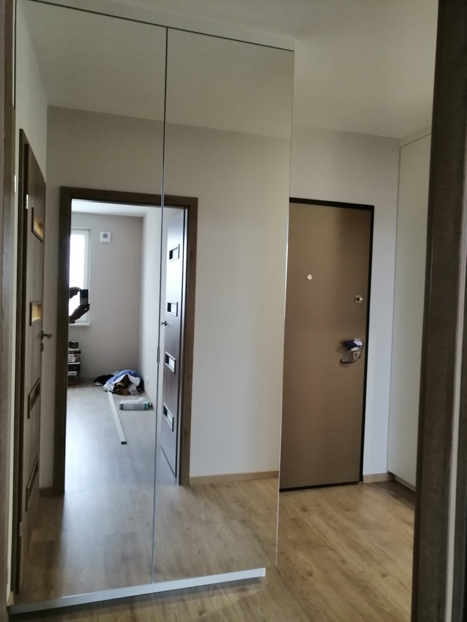 Szafa wnękowa z lustrzanymi drzwiami przesuwnymi w przedpokoju, odbijająca wnętrze mieszkania, widoczna podłoga laminowana i drzwi wejściowe.