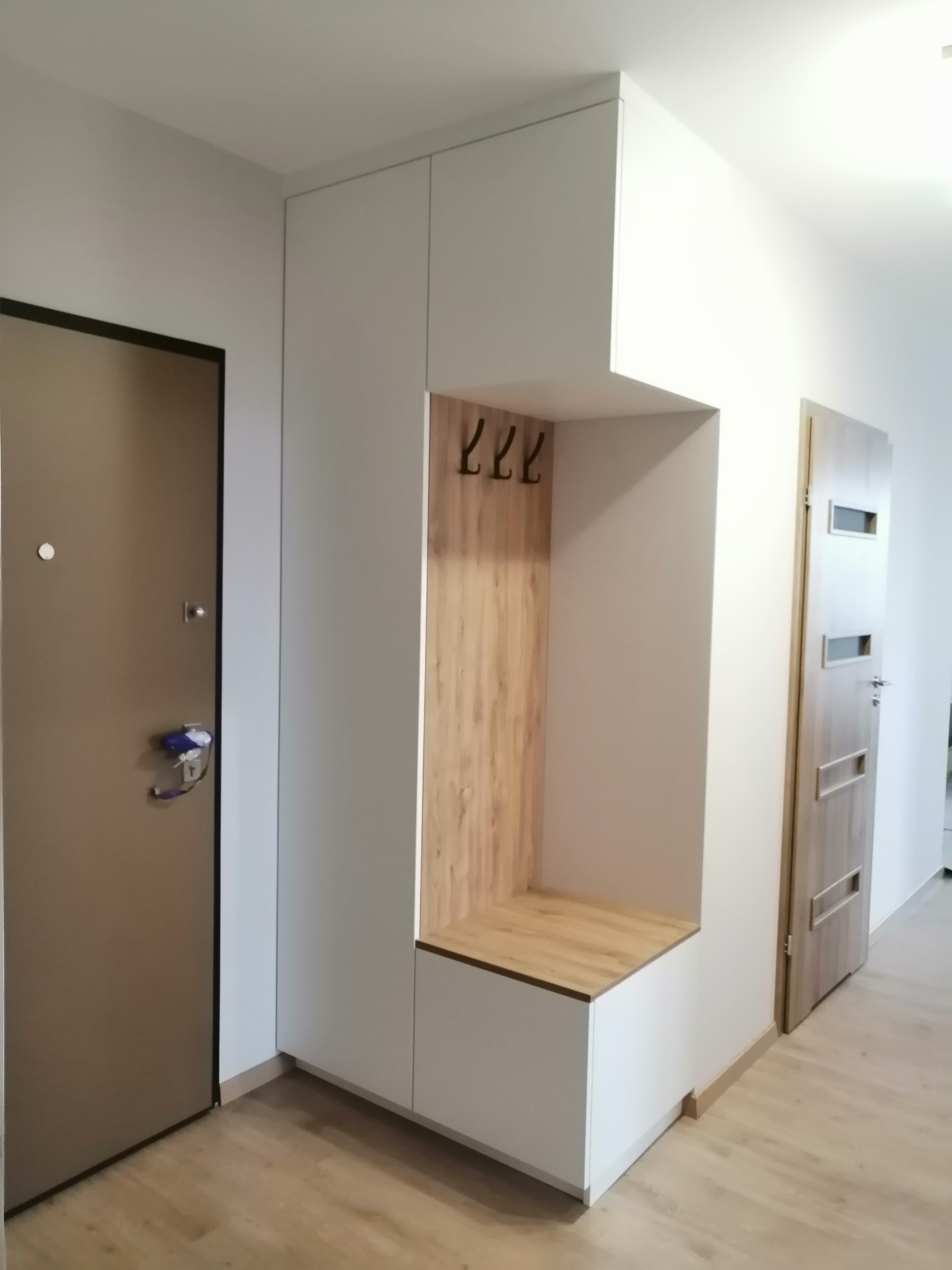 Biała szafa w przedpokoju z drewnianą wnęką na buty i wieszakami, obok drzwi wejściowych i wewnętrznych.