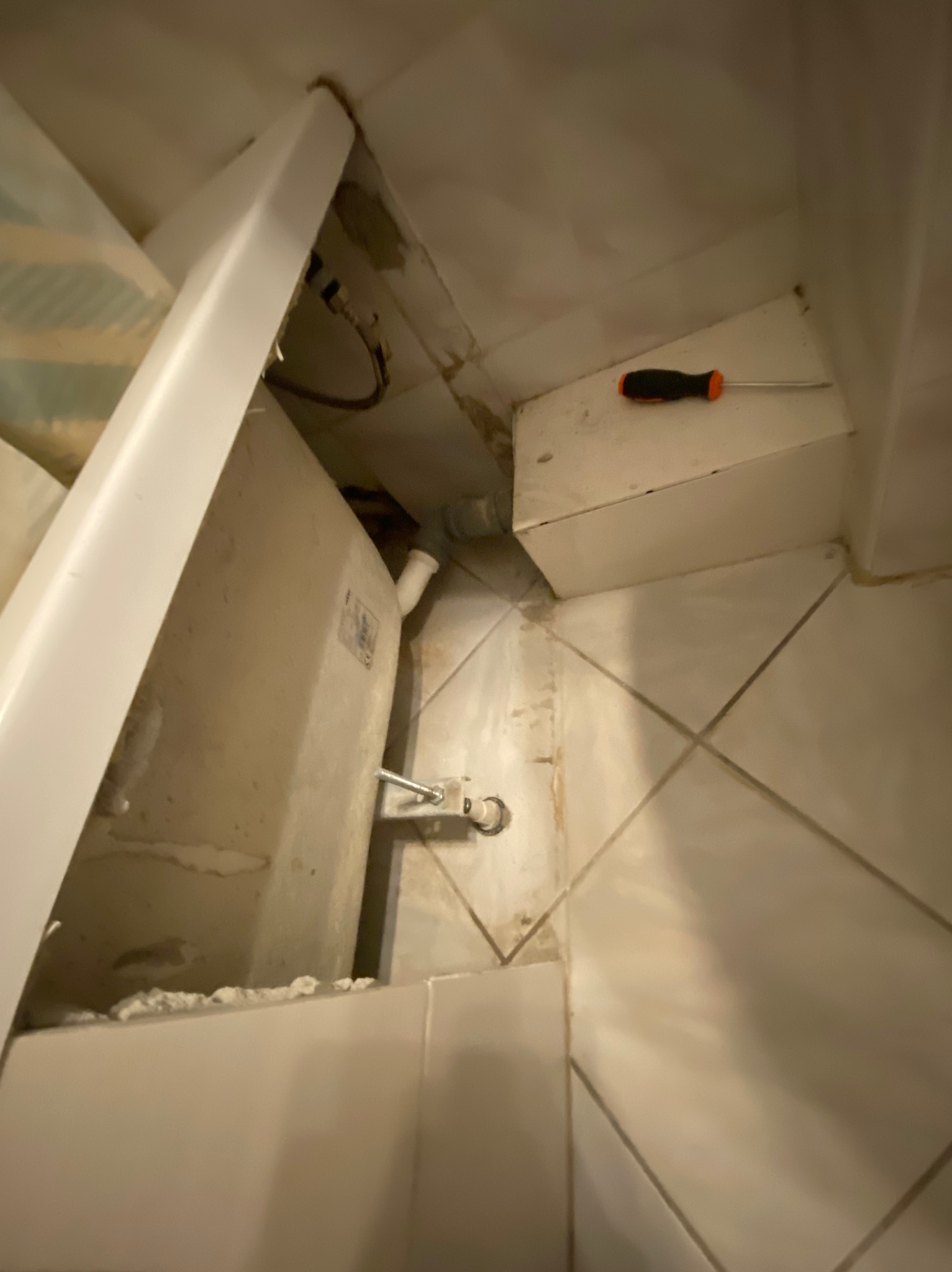 Nisko kątowe ujęcie instalacji podtynkowego zbiornika WC z widocznym odpływem, mocowaniem oraz leżącym obok śrubokrętem na tle beżowych płytek.