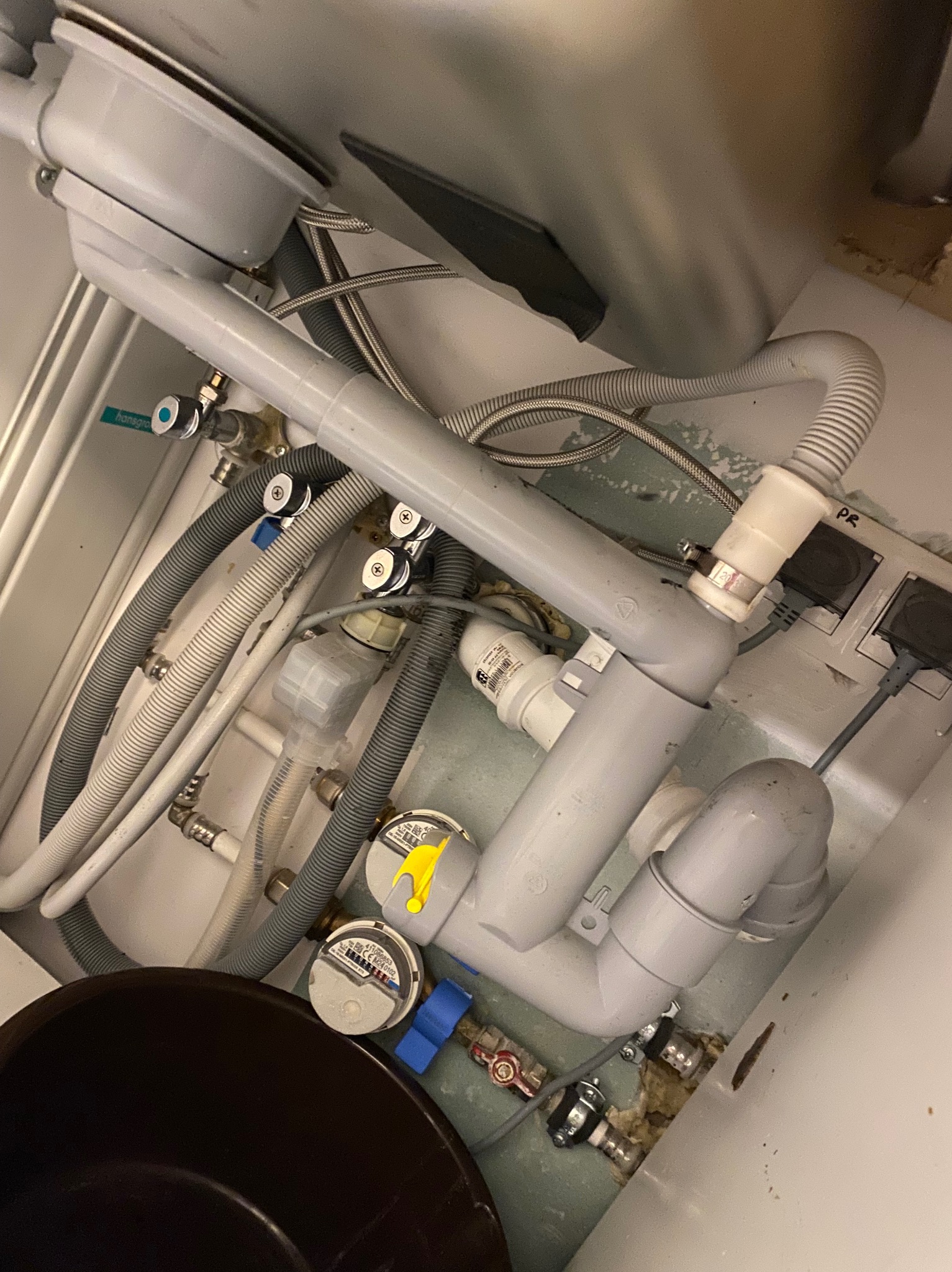 Zawiłe rury odpływowe pod zlewem z widocznymi zaworami, licznikiem wody i fragmentem czarnego wiadra