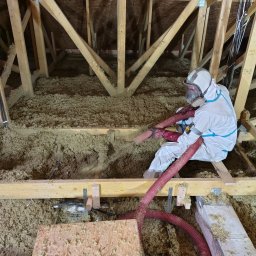 T&M Solid izolacje wdmuchiwane i wykończenia - Pracownik w kombinezonie i masce aplikuje izolację z włókien na poddaszu za pomocą węża. Widoczne drewniane elementy konstrukcyjne dachu i warstwa izolacji.