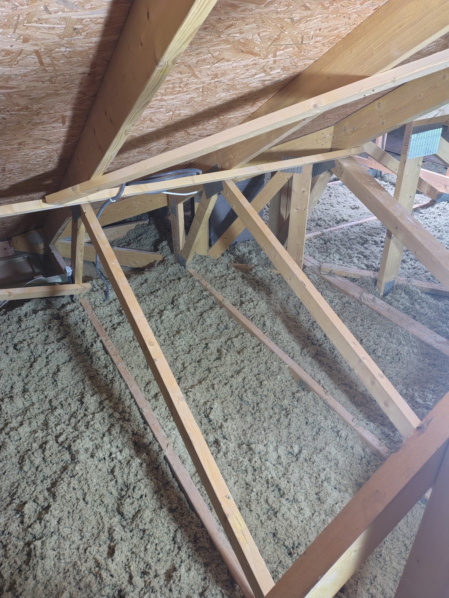 Poddasze z widoczną izolacją z wełny mineralnej między drewnianymi krokwiami. Widoczne przewody elektryczne. Ujęcie z góry, ukazujące strukturę izolacji.