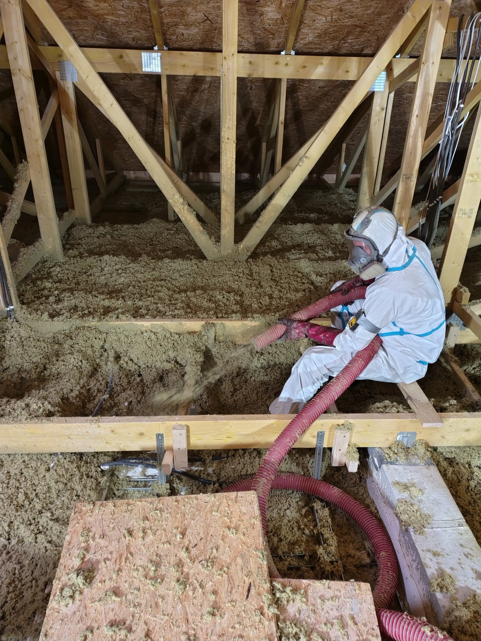 Pracownik w kombinezonie i masce aplikuje izolację z włókien na poddaszu za pomocą węża. Widoczne drewniane elementy konstrukcyjne dachu i warstwa izolacji.