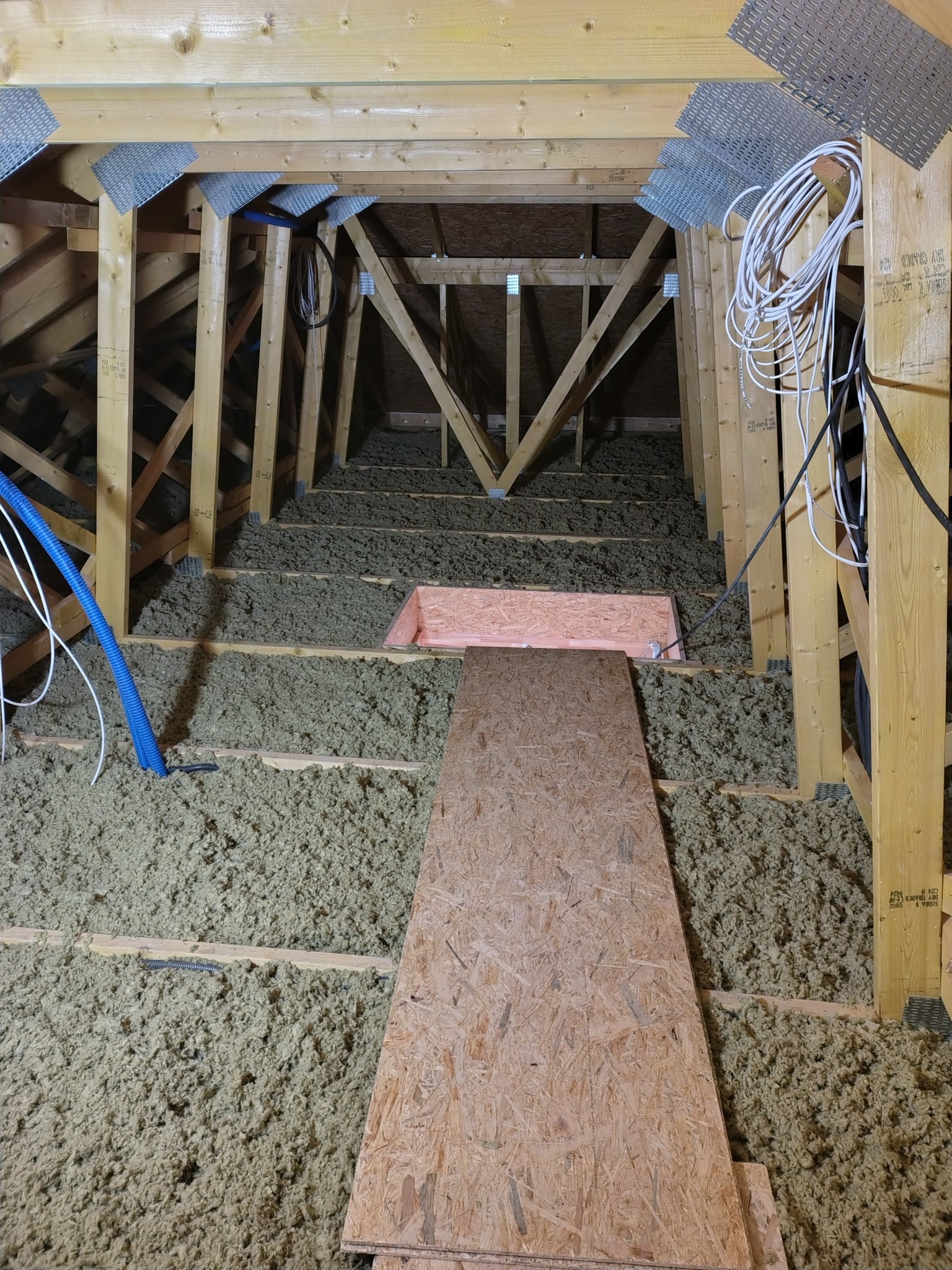 Poddasze z drewnianą konstrukcją i izolacją z wełny mineralnej, widoczne kable i płyta OSB. Ujęcie z perspektywy wnętrza poddasza, ukazujące detale izolacji.