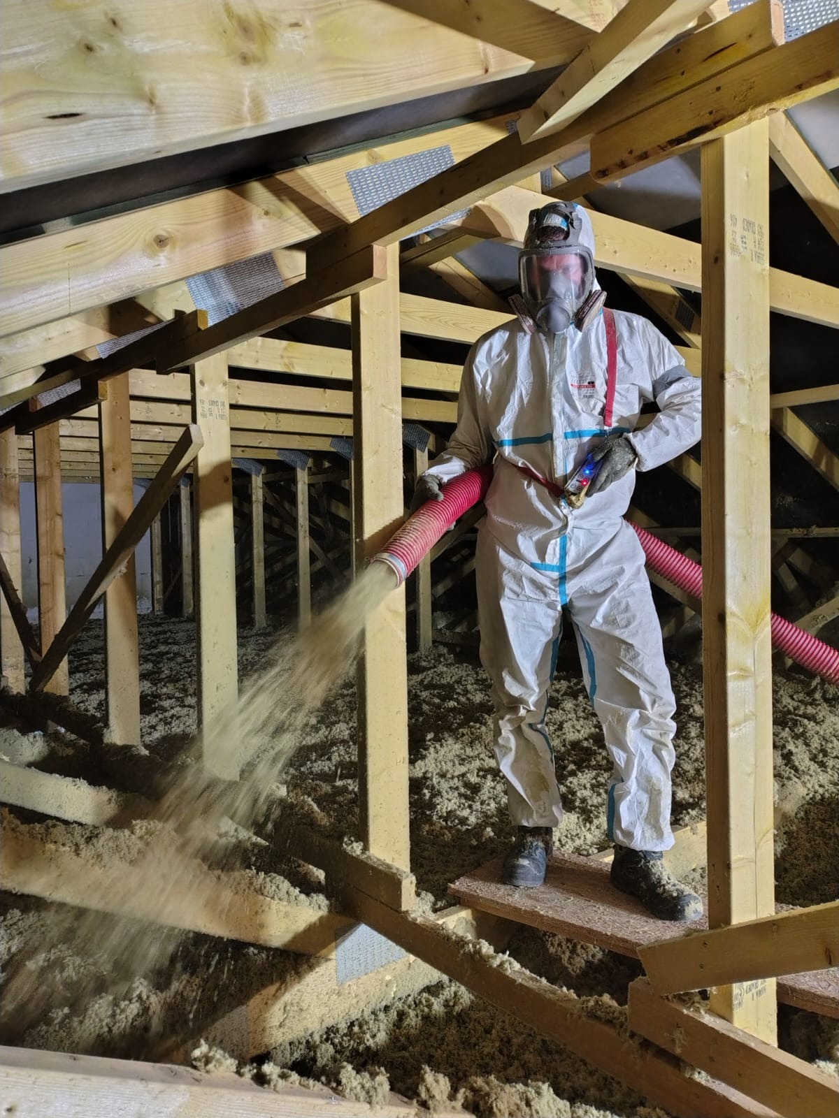 Pracownik w kombinezonie ochronnym aplikuje izolację na poddaszu w Żarach za pomocą specjalistycznego sprzętu. Widoczne drewniane elementy konstrukcji dachu.