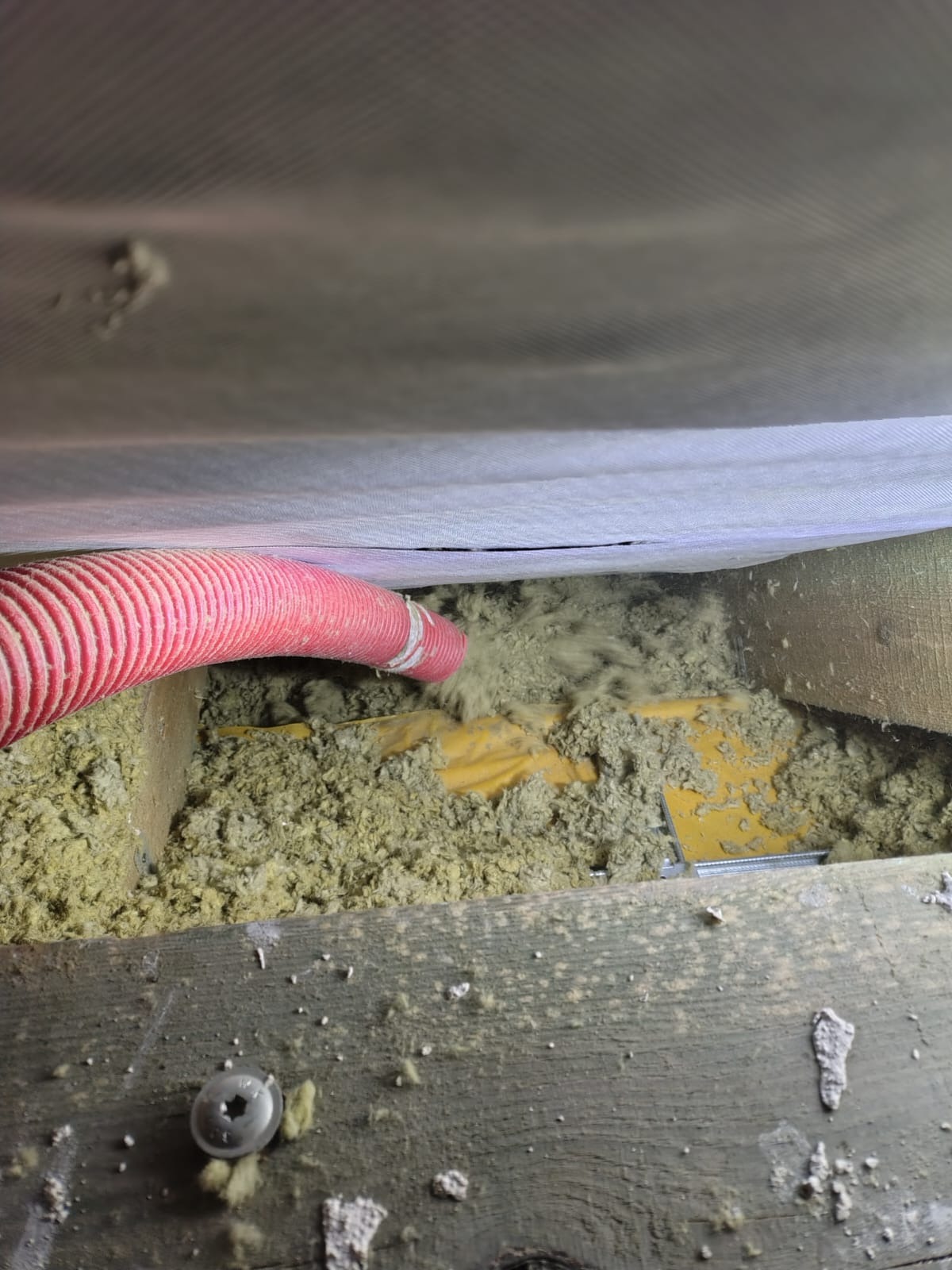 Aplikacja izolacji w przestrzeni poddasza z użyciem czerwonego węża, widoczne drewniane elementy konstrukcyjne i żółta folia izolacyjna.