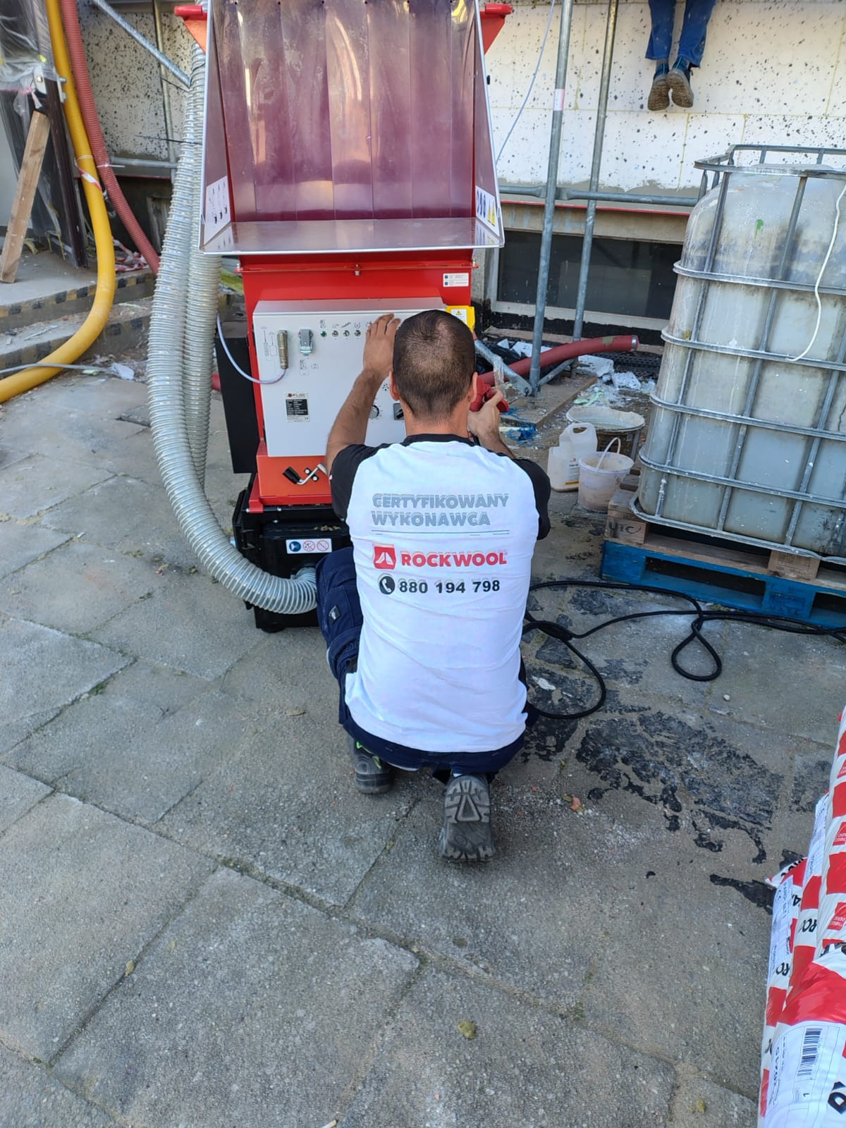 Pracownik w firmowej koszulce obsługuje maszynę do wdmuchiwania izolacji, widoczne materiały izolacyjne Rockwool i fragment ocieplanej ściany budynku.
