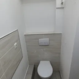 Nowoczesna toaleta w trakcie prac wykończeniowych, widoczne płytki w odcieniach szarości, podwieszana miska WC, instalacja wodna oraz narzędzia na podłodze.