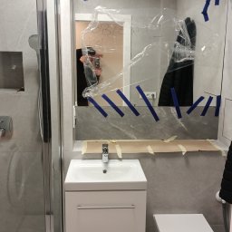 Marcin Oblak - Wykończenia łazienki w Obornikach Śląskich: Nowoczesna umywalka z szafką, toaleta wisząca i kabina prysznicowa w trakcie prac wykończeniowych, zabezpieczone lustro z niebieską taśmą. Widoczne szare...