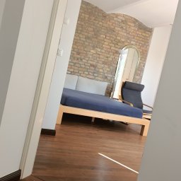 Marcin Oblak - Widok zza otwartych drzwi na sypialnię z łóżkiem na drewnianej ramie, krzesłem i dużym lustrem na tle ściany z cegły. Parkiet na podłodze.