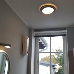 Marcin Oblak - Okrągła lampa sufitowa w łazience z widokiem na okno i grzejnik elektryczny na ścianie.