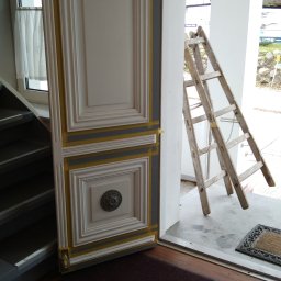 Marcin Oblak - Białe drzwi z dekoracyjnymi panelami i mosiężną gałką, zabezpieczone taśmą malarską, opierają się o ścianę obok schodów. W tle drewniana drabina i widok na budynek z dachem krytym strzechą.