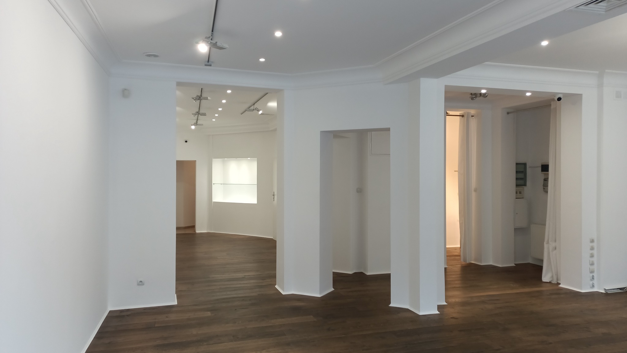 Puste, odnowione wnętrze z drewnianą podłogą, białymi ścianami, kolumnami i oświetleniem punktowym. Widoczne wejścia do innych pomieszczeń z zasłonami.