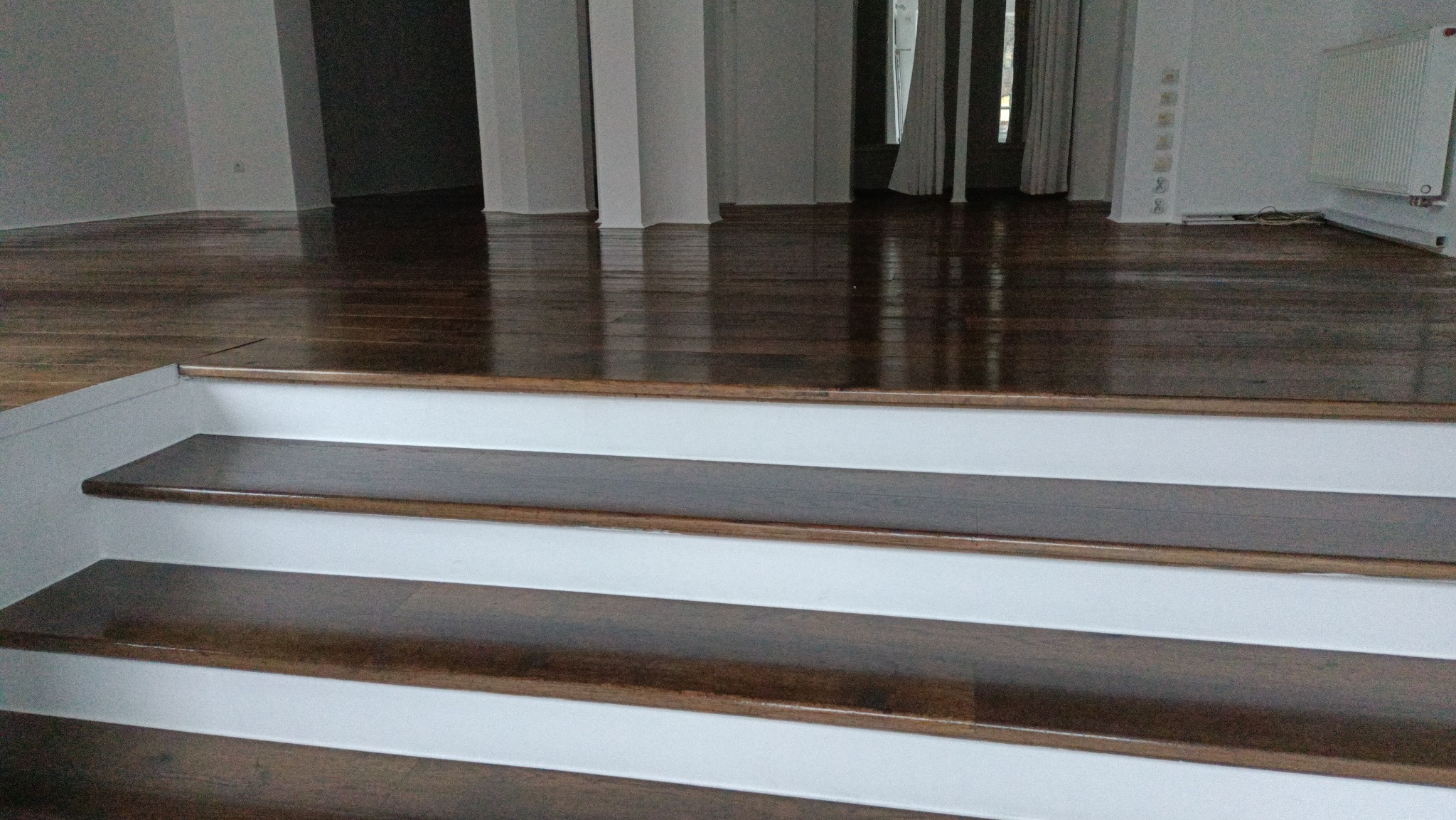 Polerowane, ciemne drewniane schody z białymi podstopnicami prowadzące do przestronnego, jasnego pomieszczenia z parkietem i kolumnami.