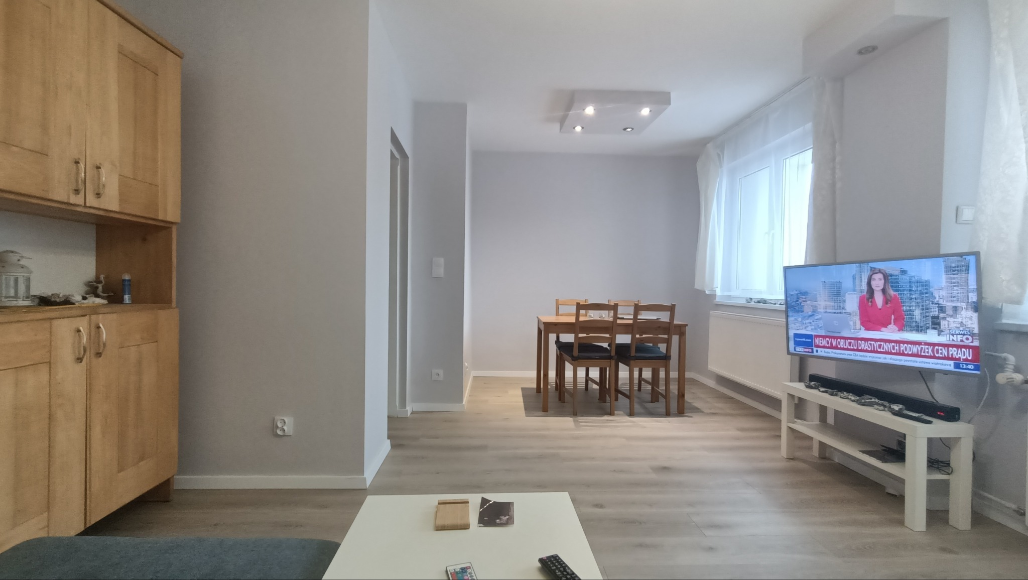 Jasne, nowocześnie urządzone wnętrze pokoju dziennego z drewnianą podłogą, szafką z jasnego drewna, stołem z krzesłami oraz telewizorem wyświetlającym wiadomości.