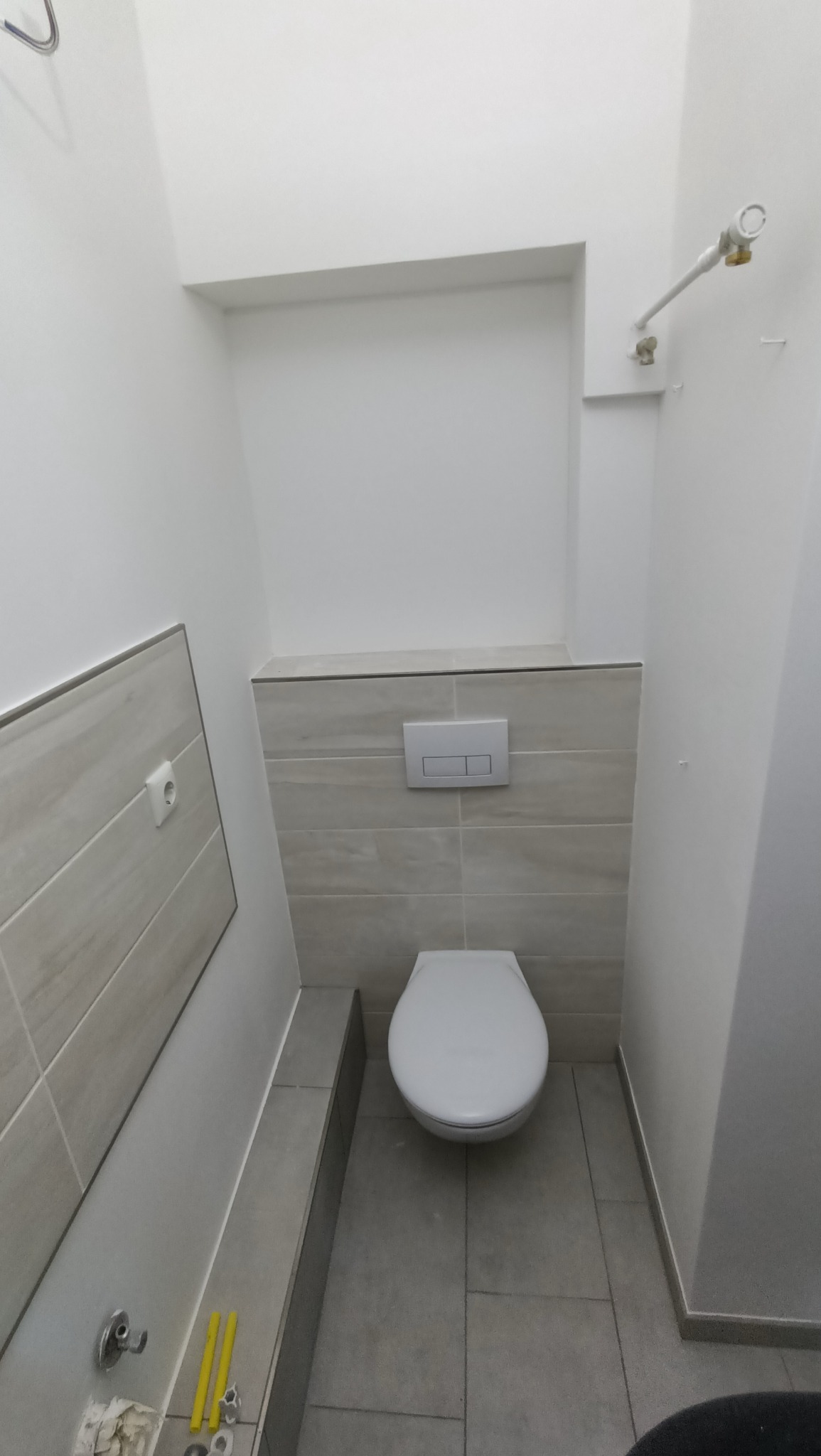 Nowoczesna toaleta w trakcie prac wykończeniowych, widoczne płytki w odcieniach szarości, podwieszana miska WC, instalacja wodna oraz narzędzia na podłodze.