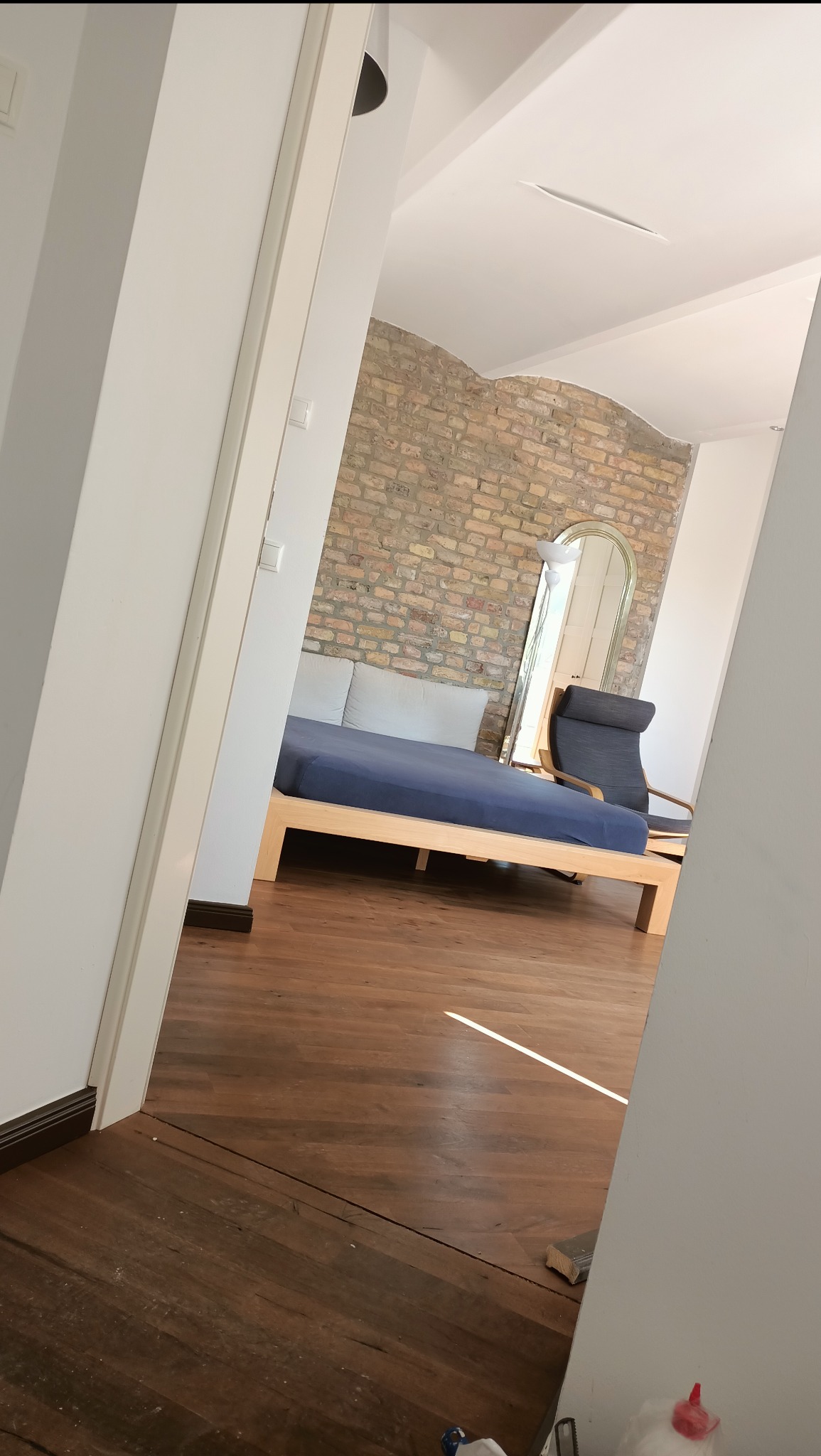 Widok zza otwartych drzwi na sypialnię z łóżkiem na drewnianej ramie, krzesłem i dużym lustrem na tle ściany z cegły. Parkiet na podłodze.