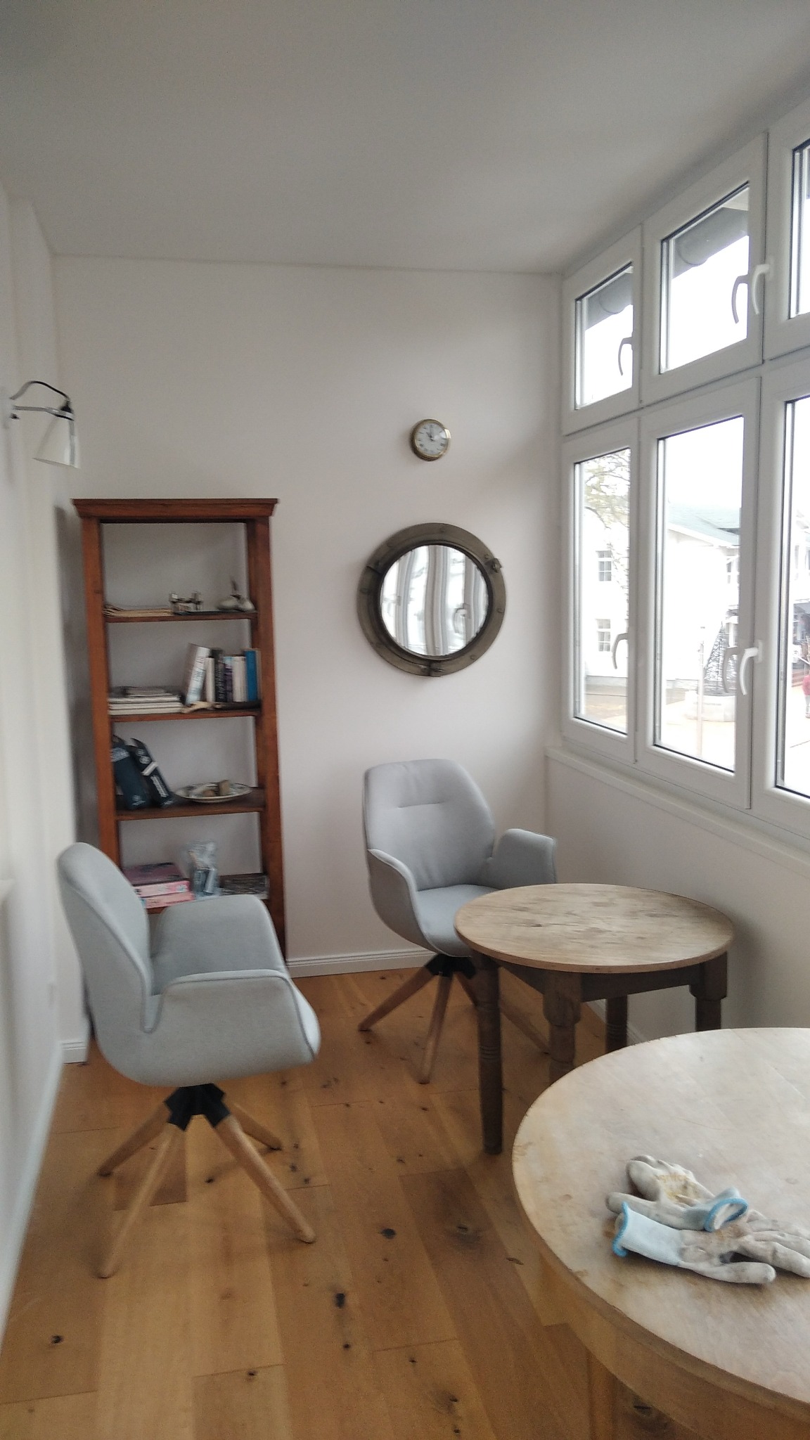 Jasne wnętrze pokoju z drewnianą podłogą, dwoma szarymi krzesłami, okrągłym stołem z parą roboczych rękawic, regałem z książkami i okrągłym lustrem na ścianie.