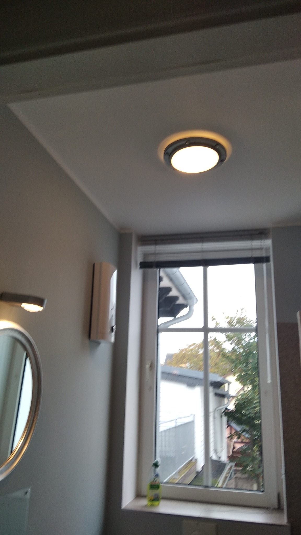 Okrągła lampa sufitowa w łazience z widokiem na okno i grzejnik elektryczny na ścianie.