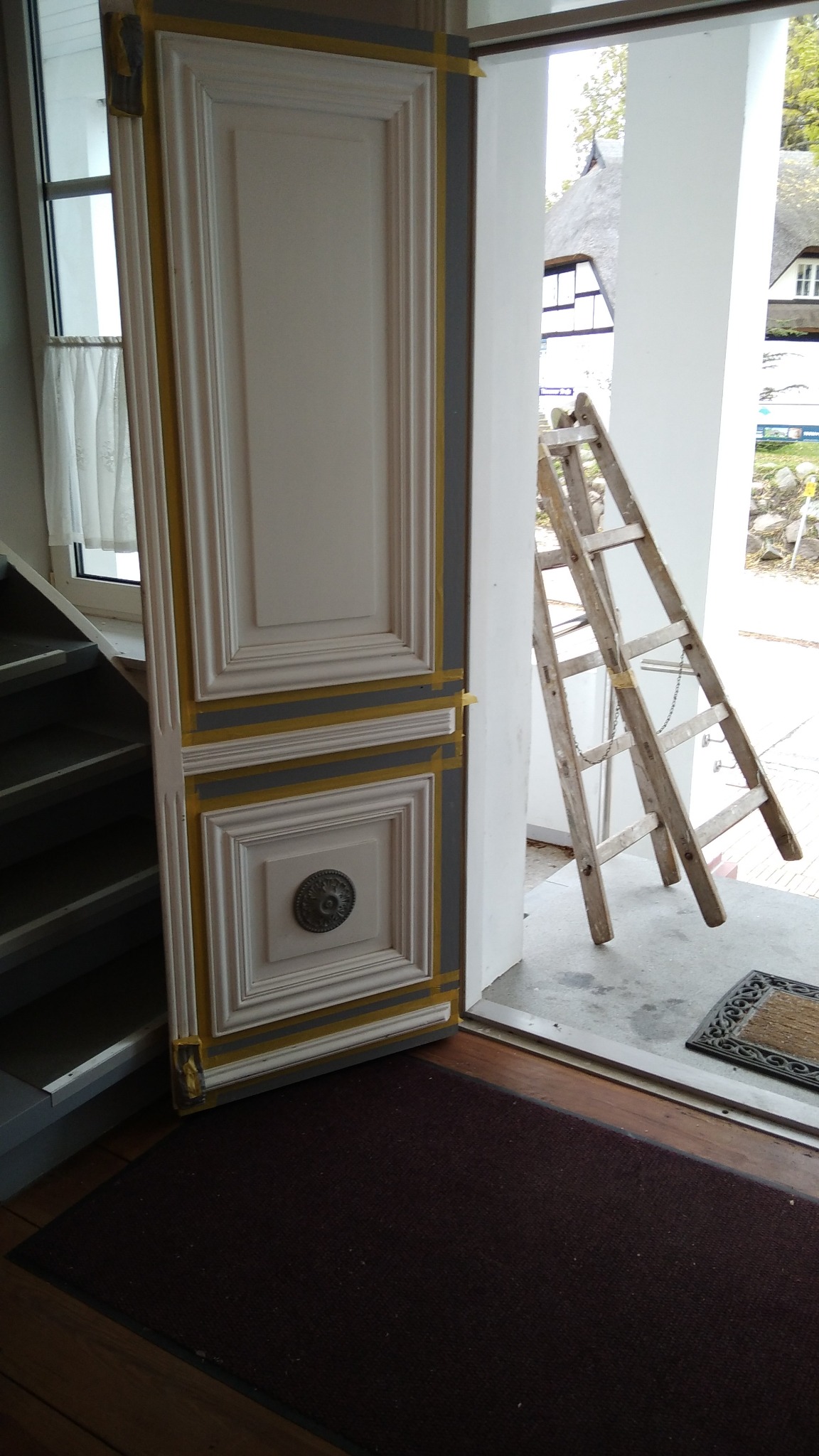 Białe drzwi z dekoracyjnymi panelami i mosiężną gałką, zabezpieczone taśmą malarską, opierają się o ścianę obok schodów. W tle drewniana drabina i widok na budynek z dachem krytym strzechą.