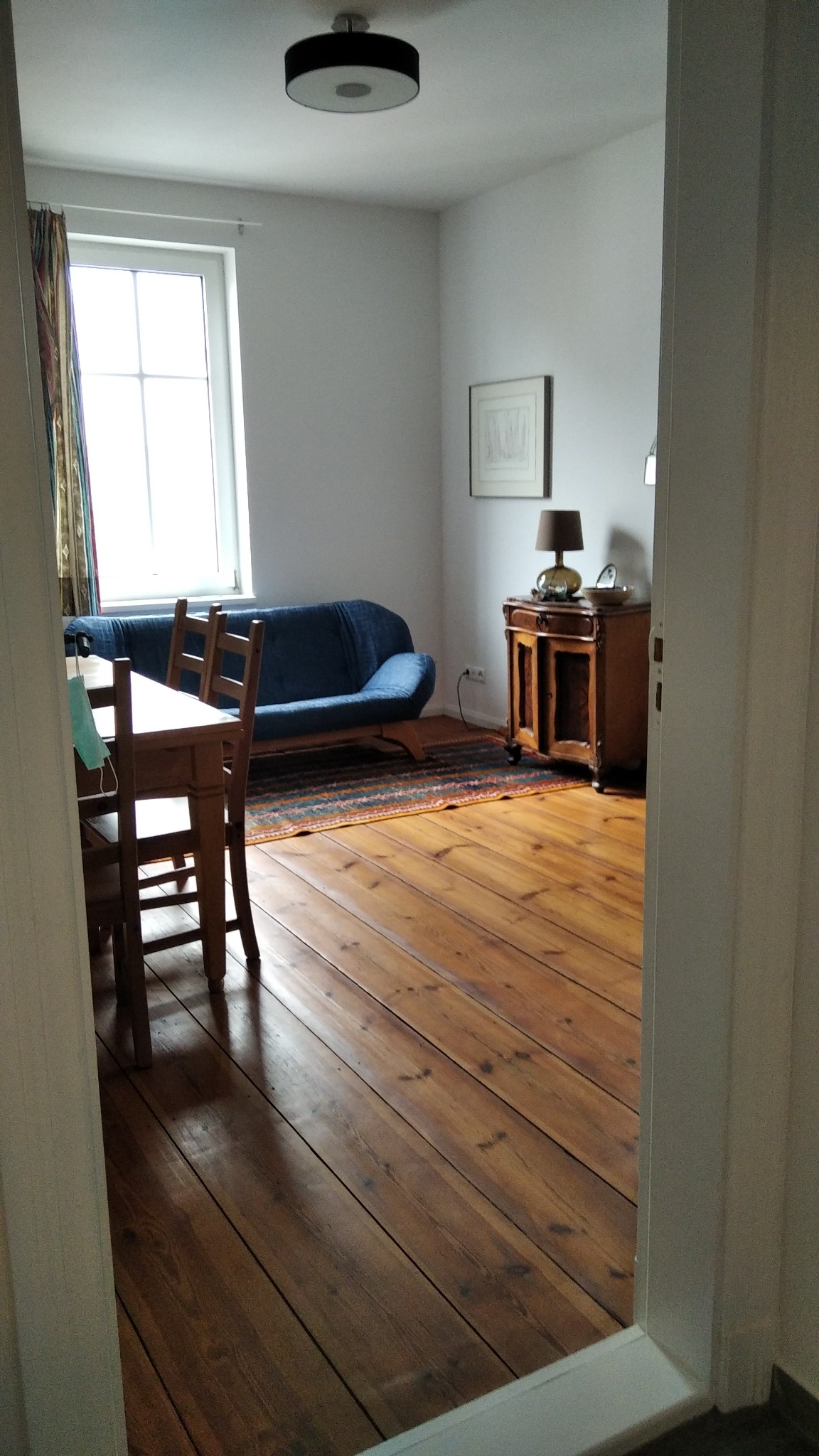 Widok z korytarza na pokój z drewnianą podłogą, niebieską sofą, stolikiem z krzesłami i lampą na komodzie, maska na krześle.