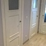 Dwa białe, klasyczne drzwi wewnętrzne z szybą w górnej części, zamontowane w szarej ścianie. Widoczna podłoga z jasnego drewna i fragment trzecich drzwi w tle.