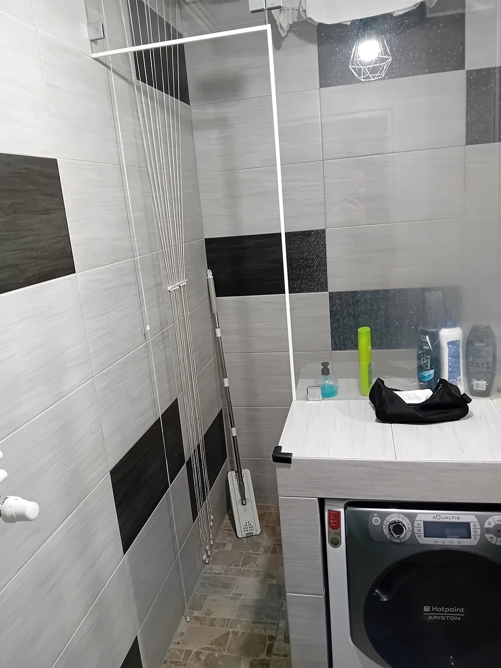 Szaro-biała łazienka z płytkami w geometryczny wzór, pralką Ariston, suszarką sufitową na pranie, mopem oraz kosmetykami na blacie.