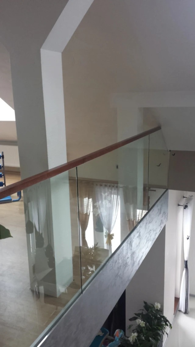 Szklana balustrada z drewnianym poręczą na antresoli, widok z góry, jasne wnętrze z białymi ścianami i zasłonami.