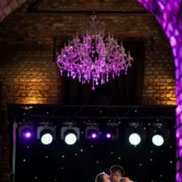 Para młoda tańczy w objęciach pod kryształowym żyrandolem, w tle ceglany łuk i oświetlenie sceniczne w odcieniach fioletu. Romantyczna atmosfera.