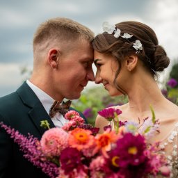 Lovegrafia MARTYNA JODŁOWSKA - Para młoda w zbliżeniu, dotykająca się nosami. Ona w sukni z kwiatami, on w ciemnozielonym garniturze i muszce. Bukiet kwiatów na pierwszym planie.