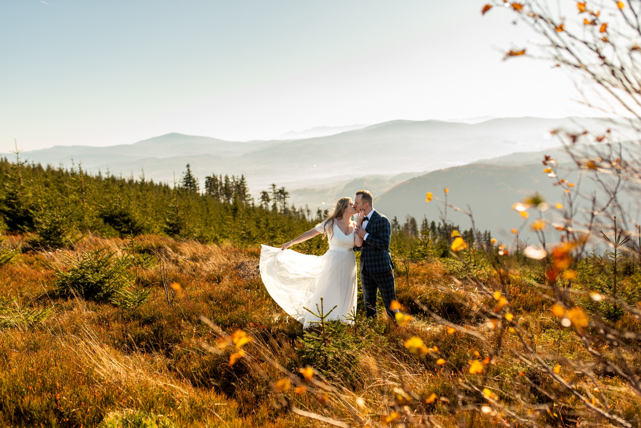 Pocałunek młodej pary w plenerze górskim, panna młoda w zwiewnej sukni, pan młody w kraciastej marynarce, jesienne trawy i drzewa, mgliste góry w tle.