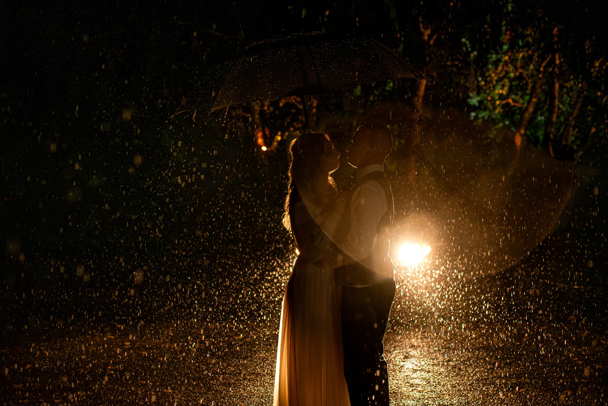 Para młoda obejmuje się pod parasolem w strugach deszczu, oświetlona od tyłu światłem samochodowym, tworząc romantyczną, choć nieco mroczną atmosferę.