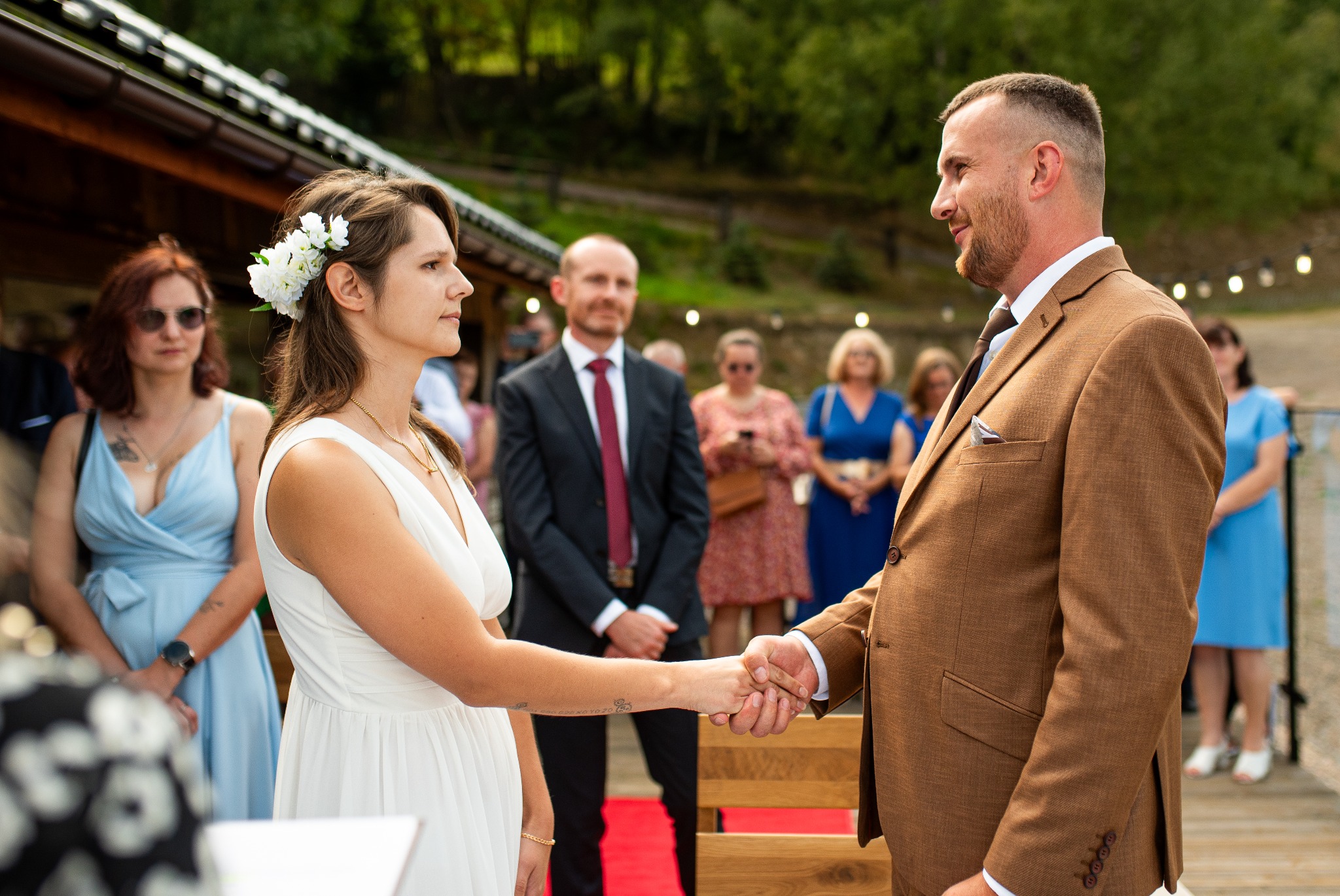 Panna młoda z kwiatami we włosach i pan młody w brązowym garniturze trzymają się za ręce podczas ceremonii ślubnej w Zatorze, z gośćmi w tle.