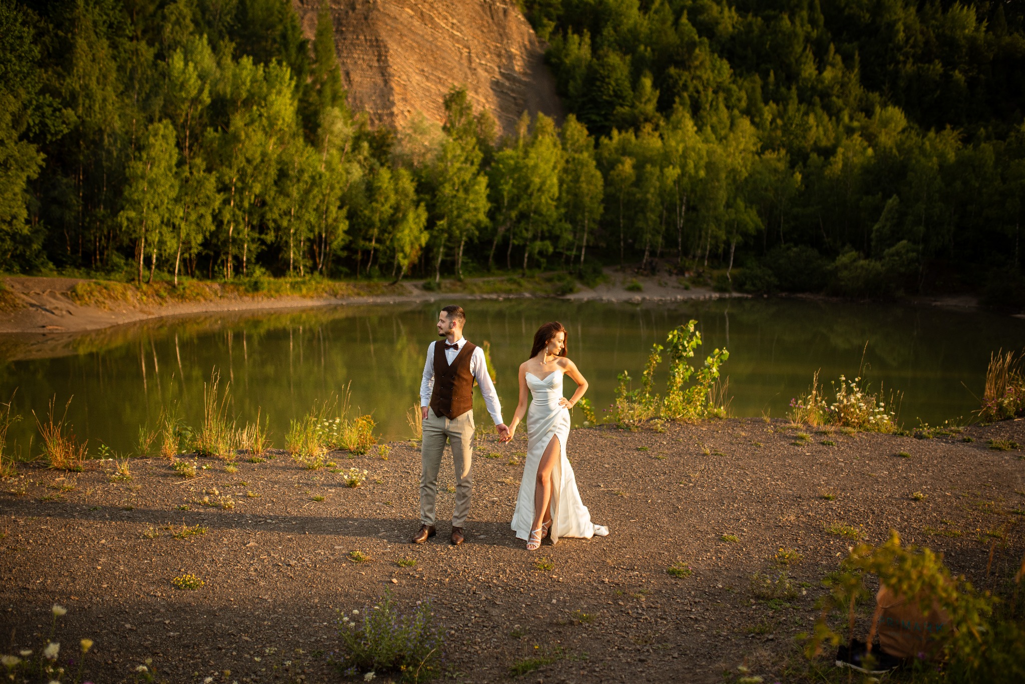 Para młoda trzymająca się za ręce nad brzegiem jeziora, on w brązowej kamizelce i muszce, ona w białej sukni z rozcięciem, w tle zielone drzewa i skalista ściana.