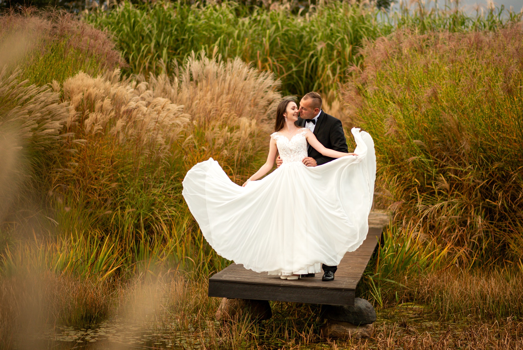 Uśmiechnięta para młoda na drewnianym pomoście wśród trzcin; panna młoda w białej sukni z rozłożystym dołem, pan młody w czarnym smokingu.
