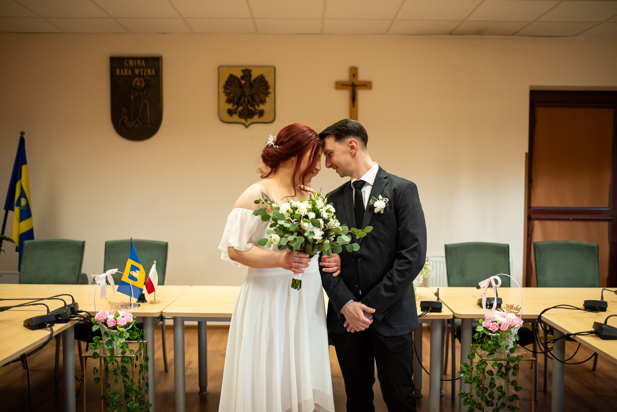 Para młoda w urzędzie stanu cywilnego, wzajemnie pochyleni głowami, panna młoda trzyma bukiet z białych kwiatów i zielonych liści eukaliptusa, w tle godło Polski i krzyż.