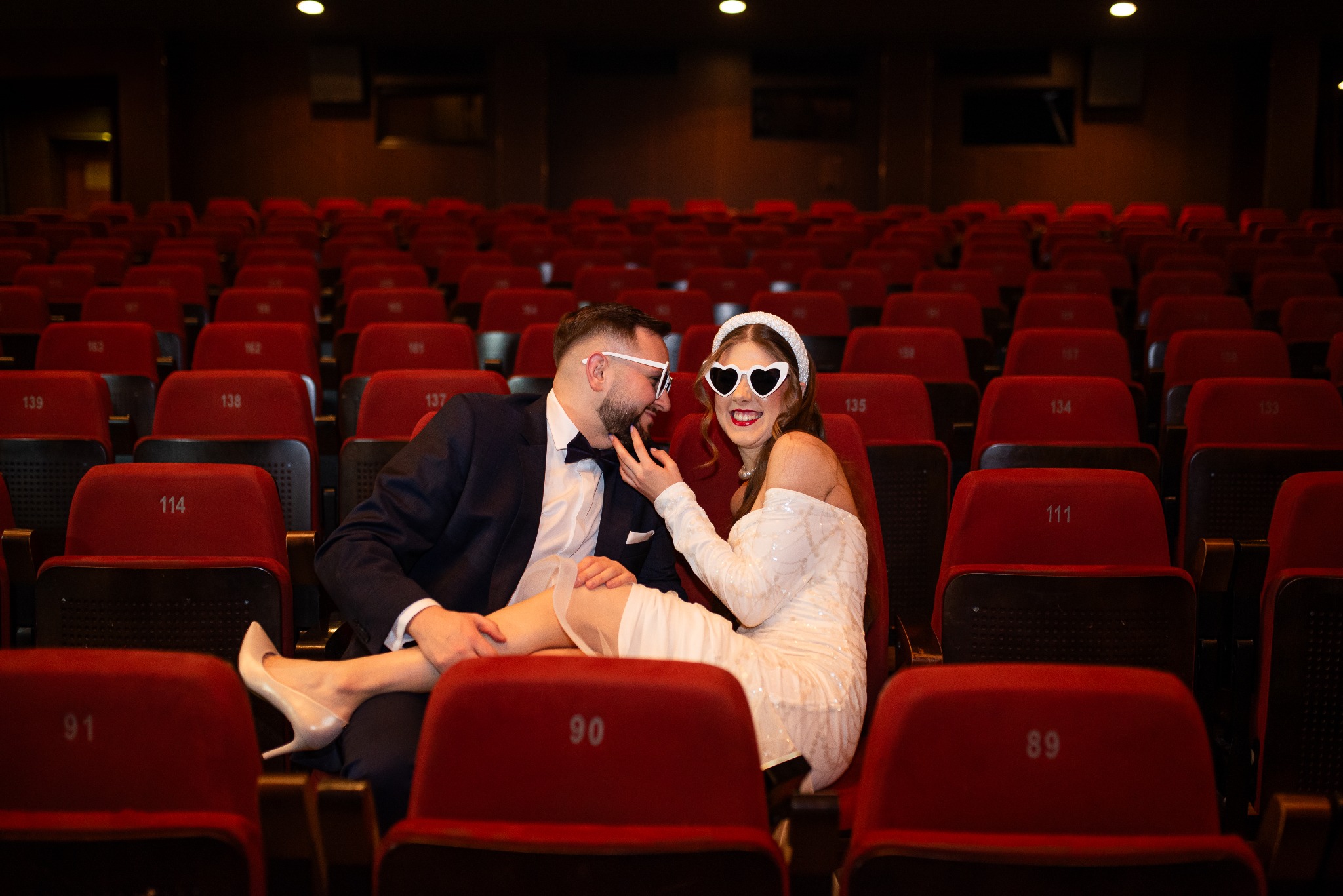 Para w kinie, ona w białej sukience i okularach w kształcie serc, on w garniturze, siedzą na czerwonych fotelach, uśmiechnięci, sesja narzeczeńska, nietypowa lokalizacja.