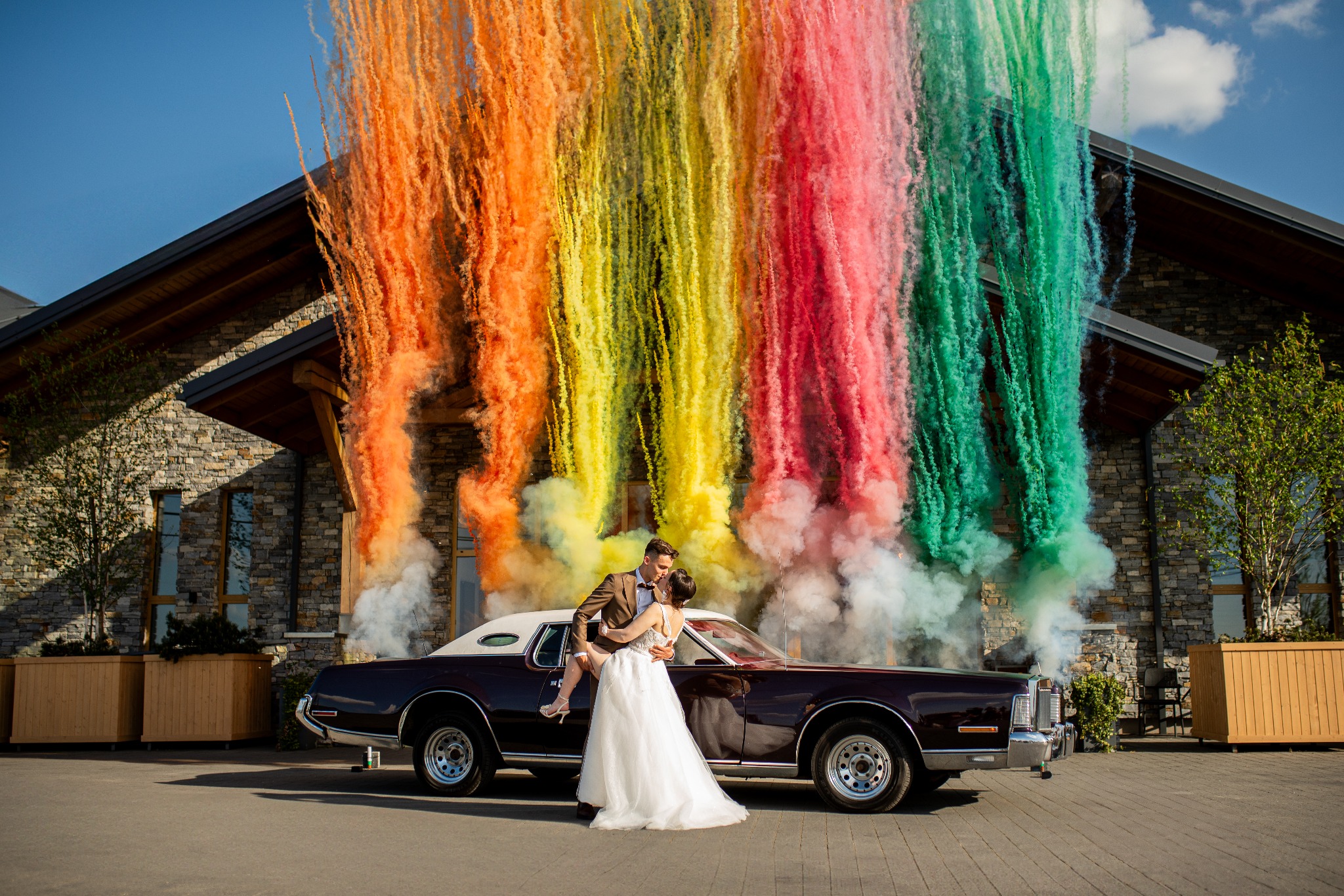 Para młoda w objęciach na tle klasycznego samochodu i kolorowych dymów, budynek w tle. Wyjątkowa sesja plenerowa z efektem specjalnym.