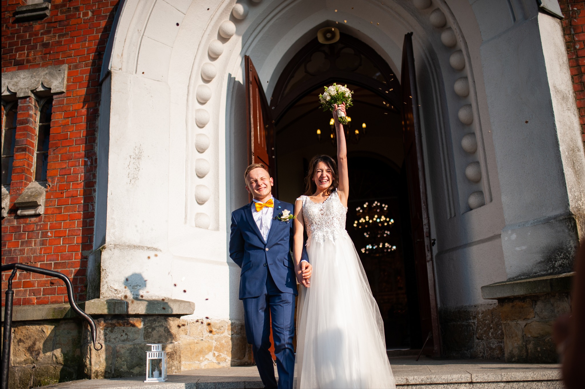 Uśmiechnięci nowożeńcy wychodzą z kościoła, panna młoda z bukietem w górze, konfetti w powietrzu, błękitny garnitur pana młodego z żółtą muchą.