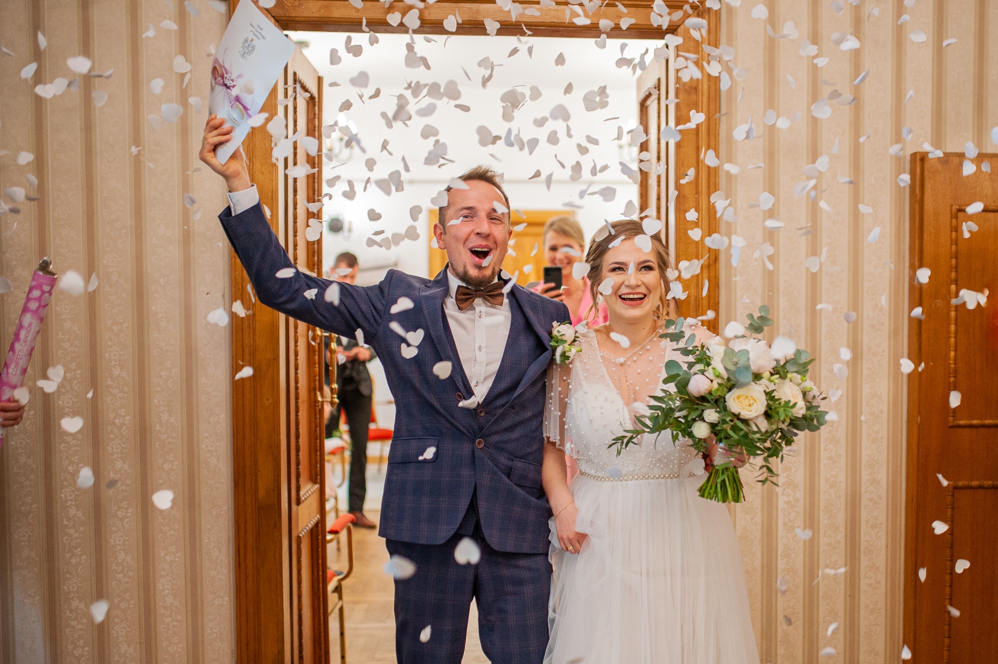 Uśmiechnięta para młoda wychodzi z sali podczas ceremonii ślubnej, obsypywana konfetti w kształcie serc. Pan młody w granatowym garniturze, panna młoda z bukietem.